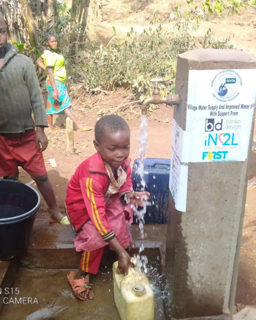A child in a red tracksuit crouching at a new concrete water standpipe in a Cameroon village, filling a yellow jerrycan from the tap while another child and a woman look on