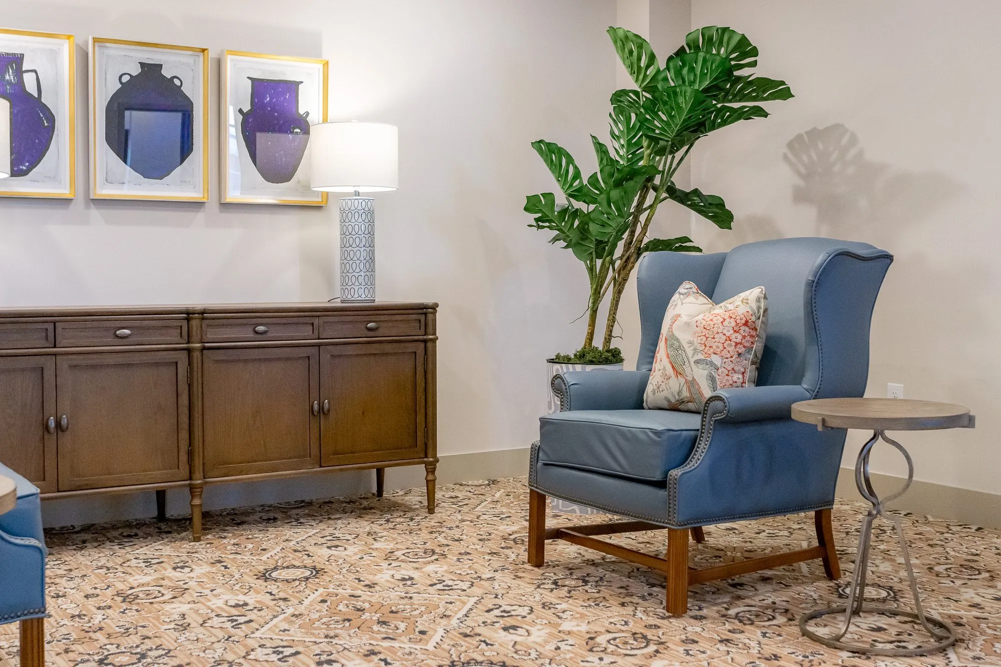Senior living interior detail: blue leather wing chair with a floral pillow beside a walnut sideboard under vase prints