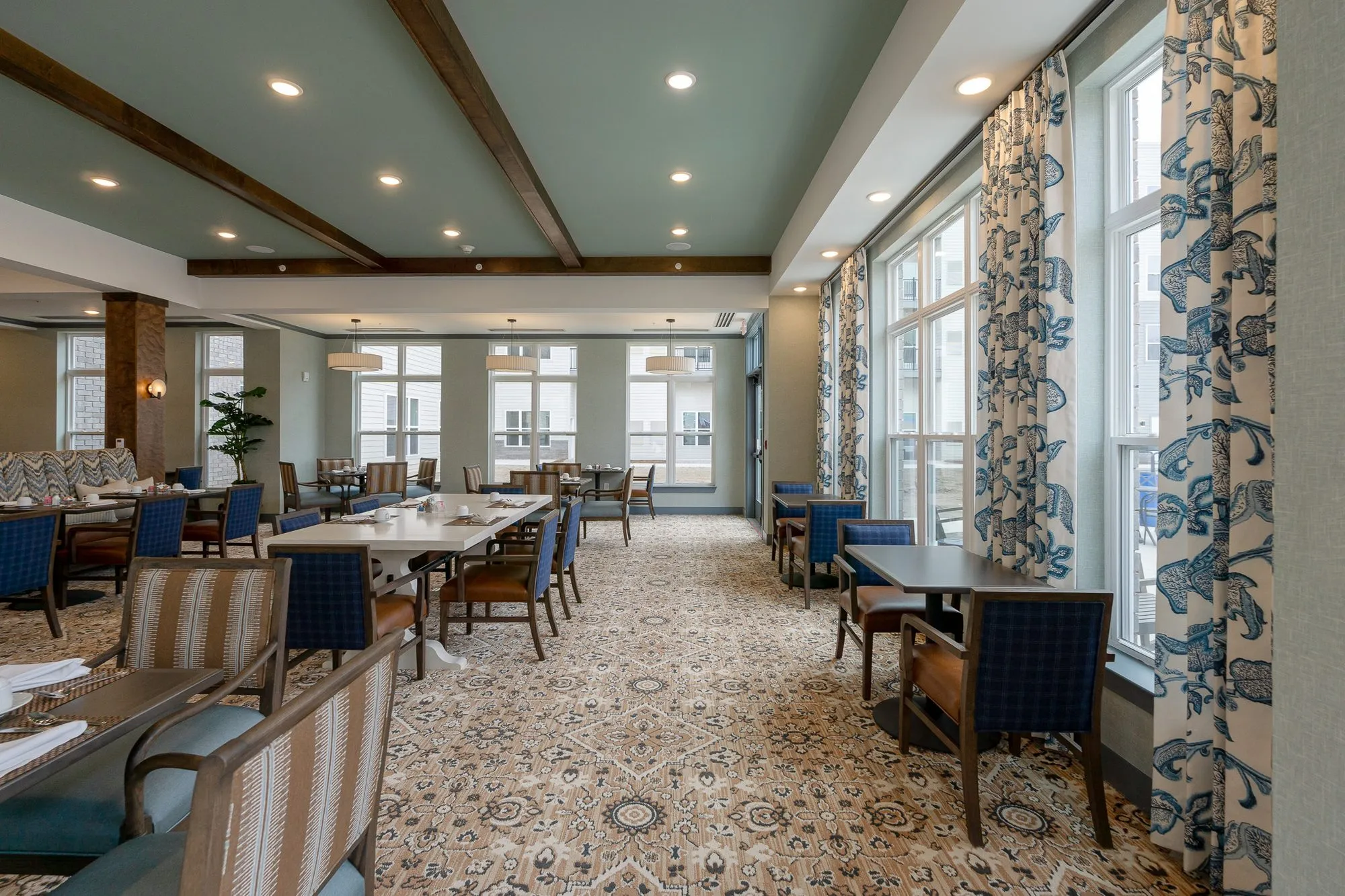 Senior living dining room under dark timber beams, blue chairs at tables and blue patterned curtains along tall windows