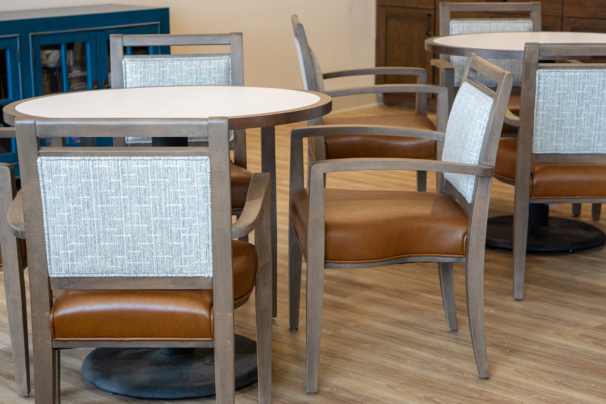 Senior living interior detail: grey wood dining chairs with tweed backs and tan leather seats at round tables