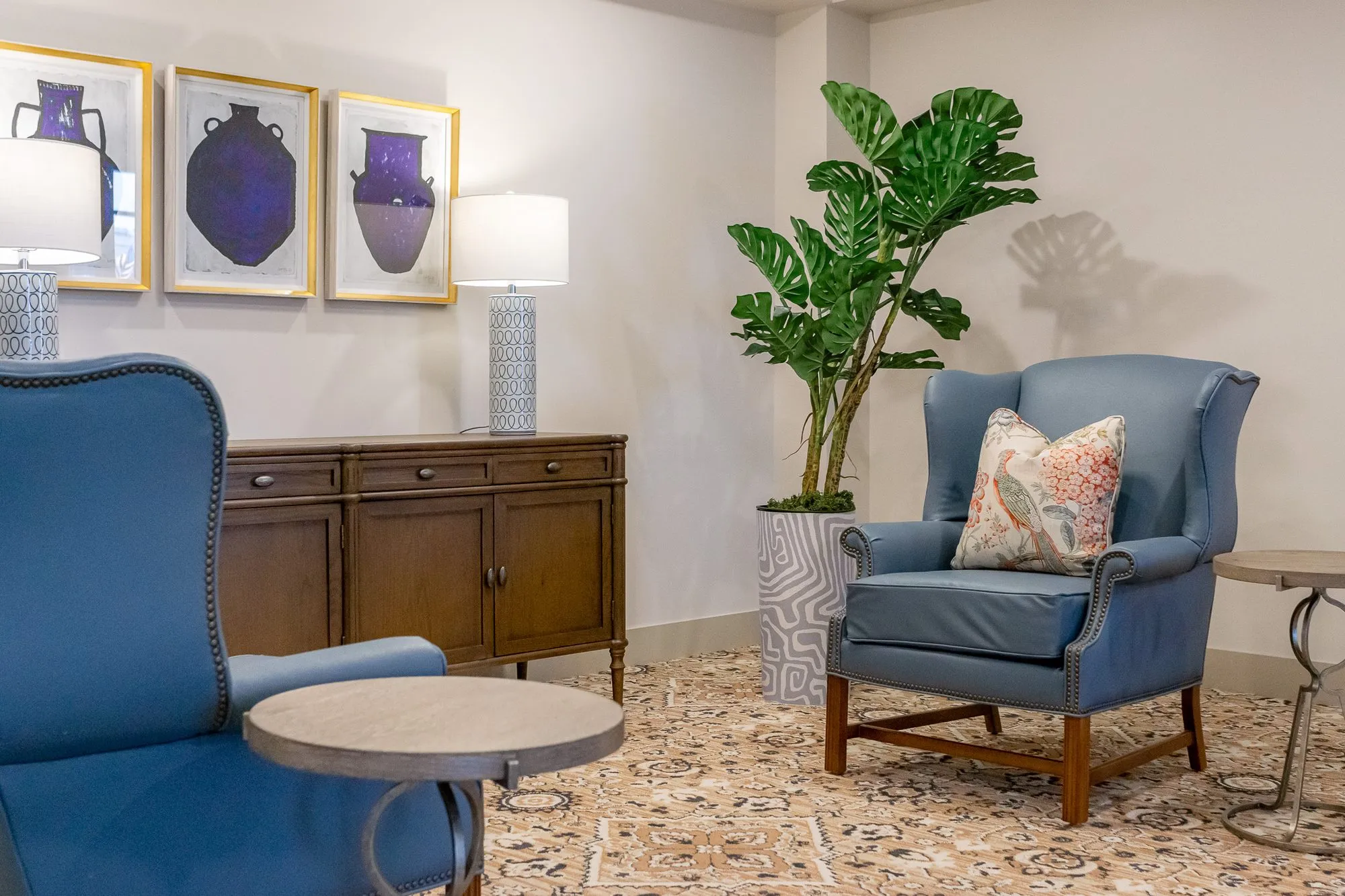 Senior living sitting area with blue leather wing chairs and a walnut sideboard with ceramic lamps and framed vase prints