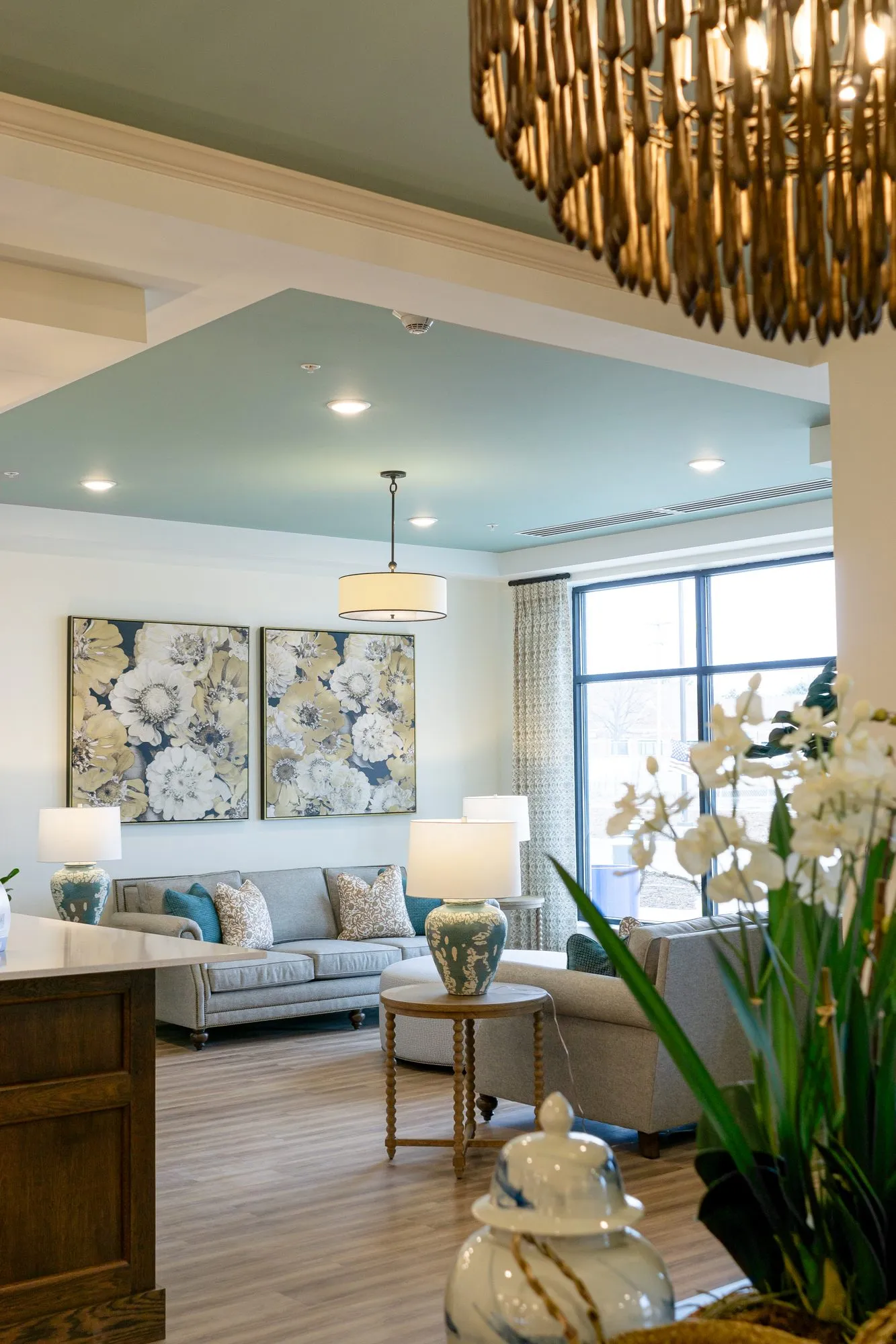 Senior living lounge with a grey sofa below two floral canvases, seen under a wood bead chandelier, orchids in front