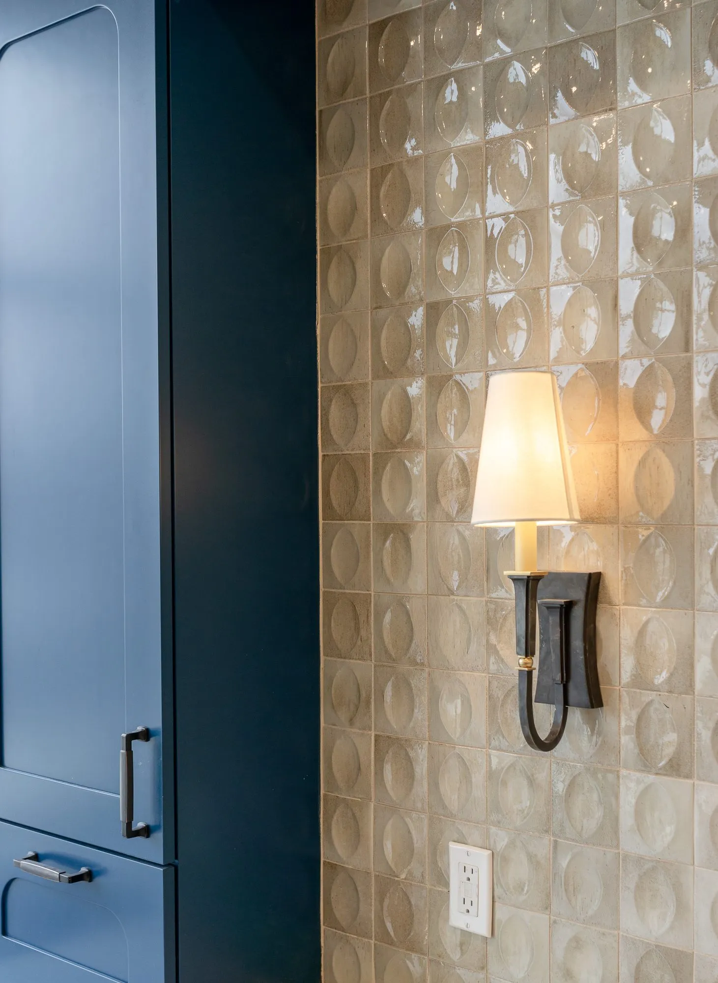 Senior living interior detail: bronze sconce with a cream shade on glossy lens pattern tile beside navy cabinetry