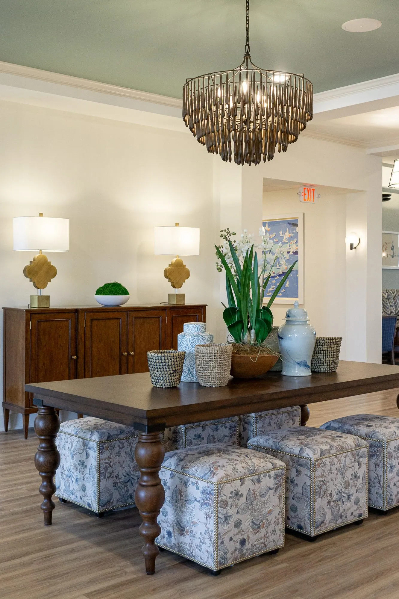 Senior living grand corridor with a walnut table and floral cube ottomans under a wood bead chandelier, a sideboard and lamps
