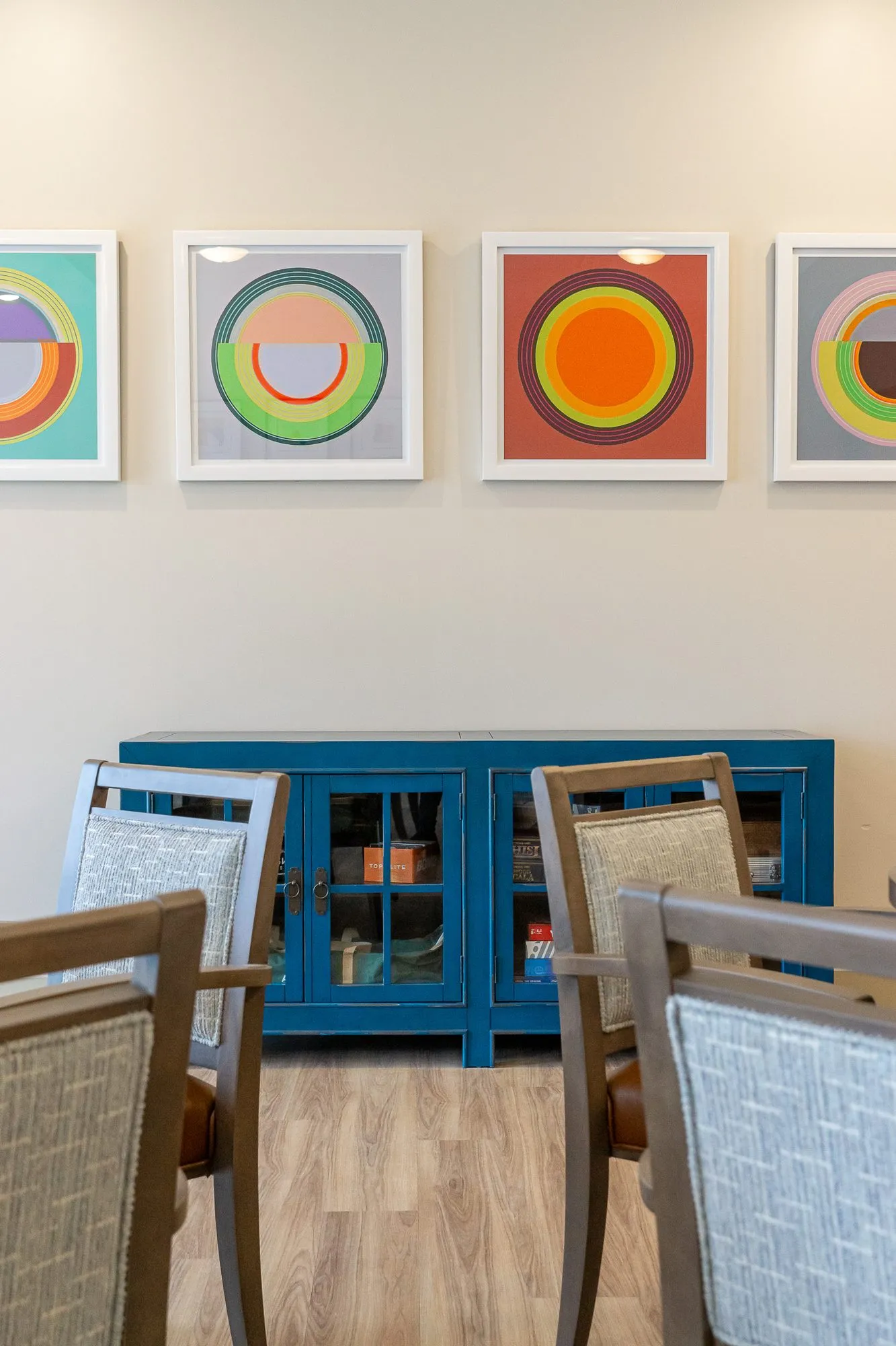 Senior living interior detail: four framed concentric circle prints above a blue glass front cabinet, grey wood chairs