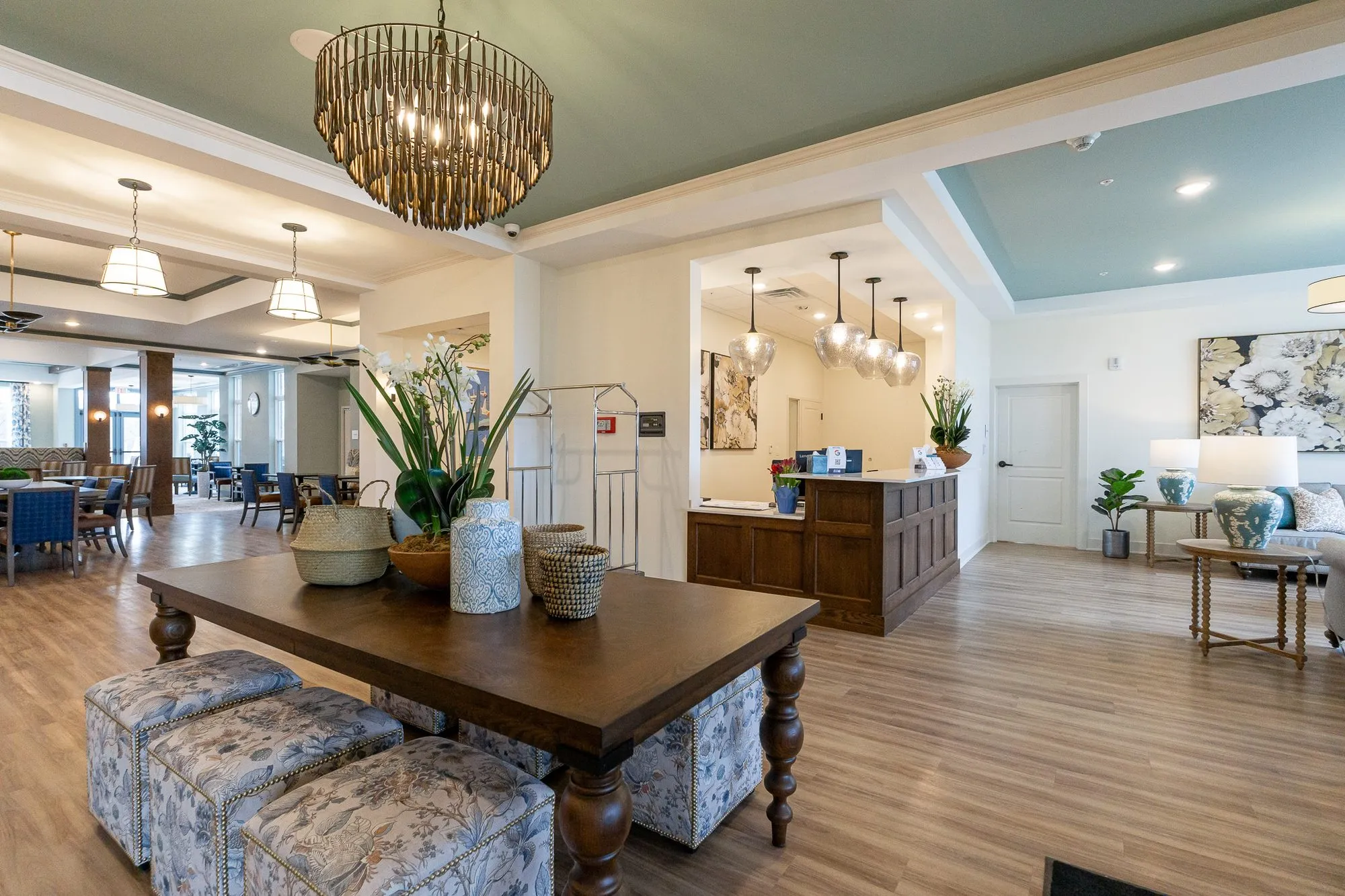 Senior living grand corridor with a walnut table and floral ottomans under a bead chandelier, concierge desk beyond