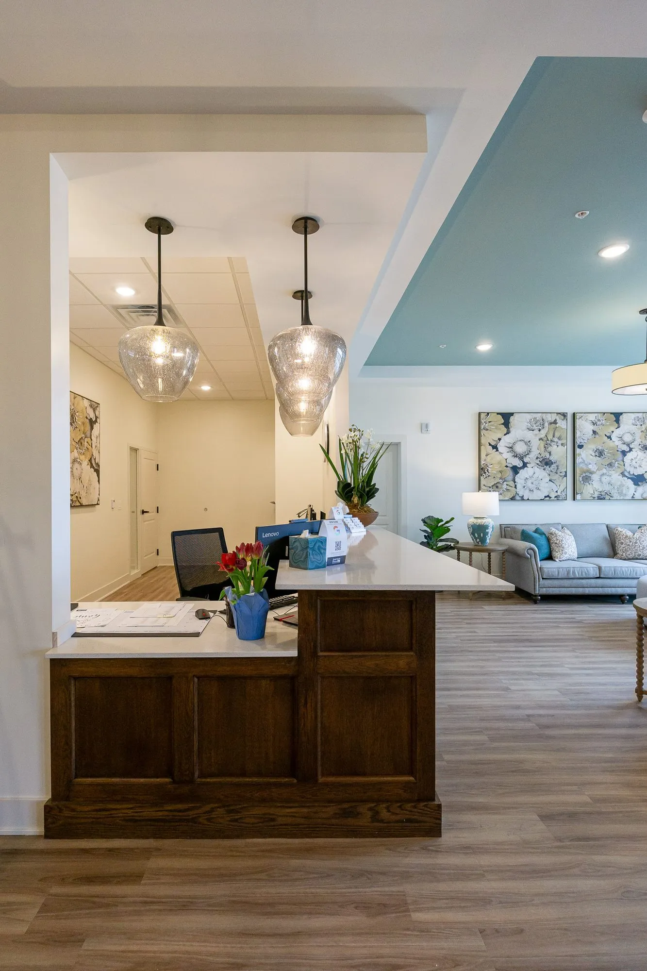 Senior living concierge desk in walnut with a white counter under glass pendants, the lounge and floral canvases beyond
