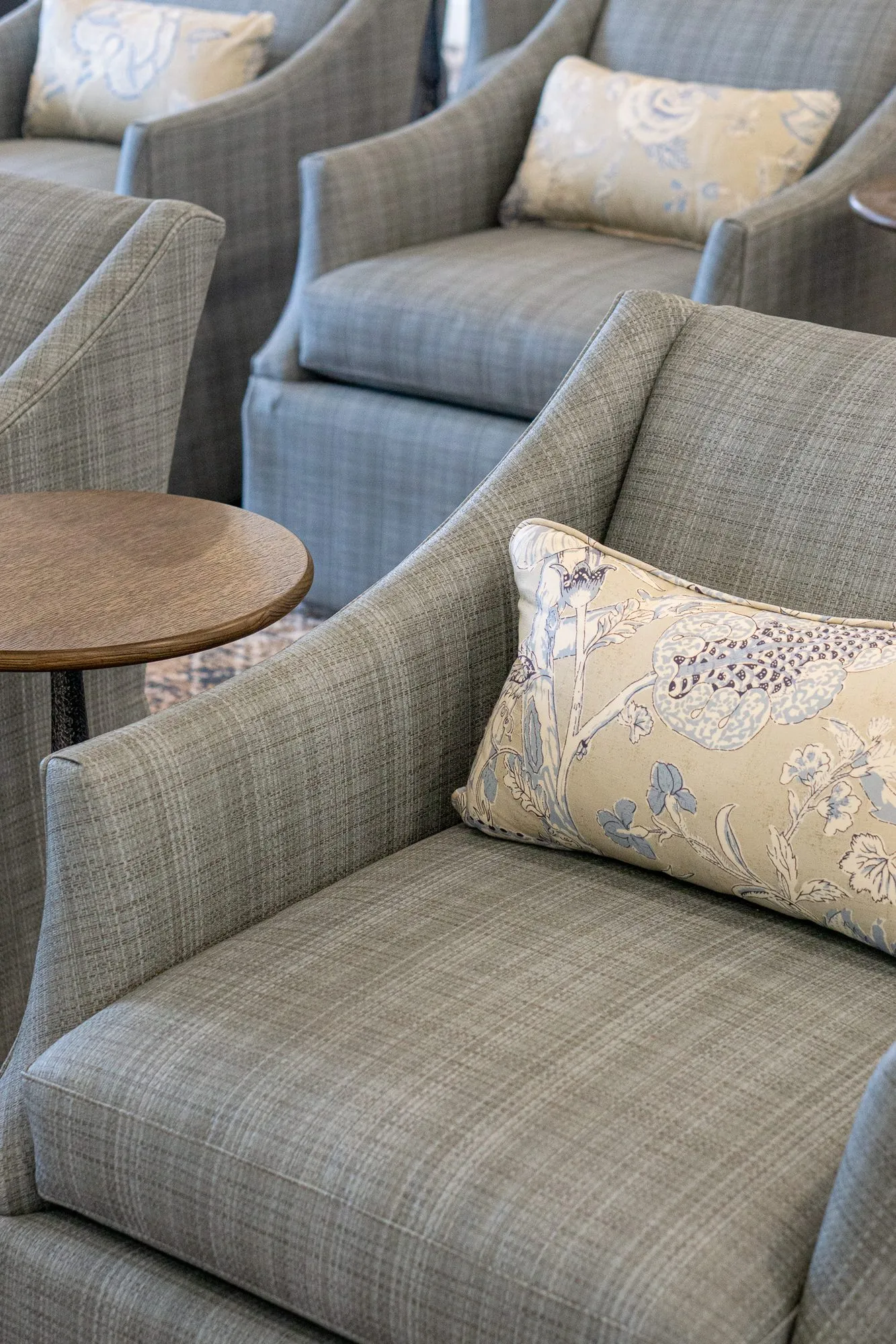 Senior living interior detail: grey plaid armchair with a floral lumbar pillow, more armchairs and a drink table behind