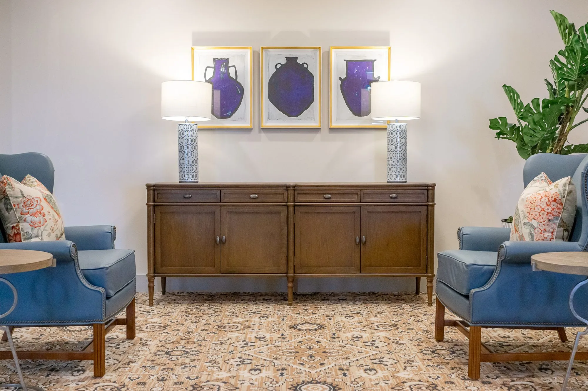 Senior living interior detail: walnut sideboard with ceramic lamps under framed vase prints between blue leather wing chairs