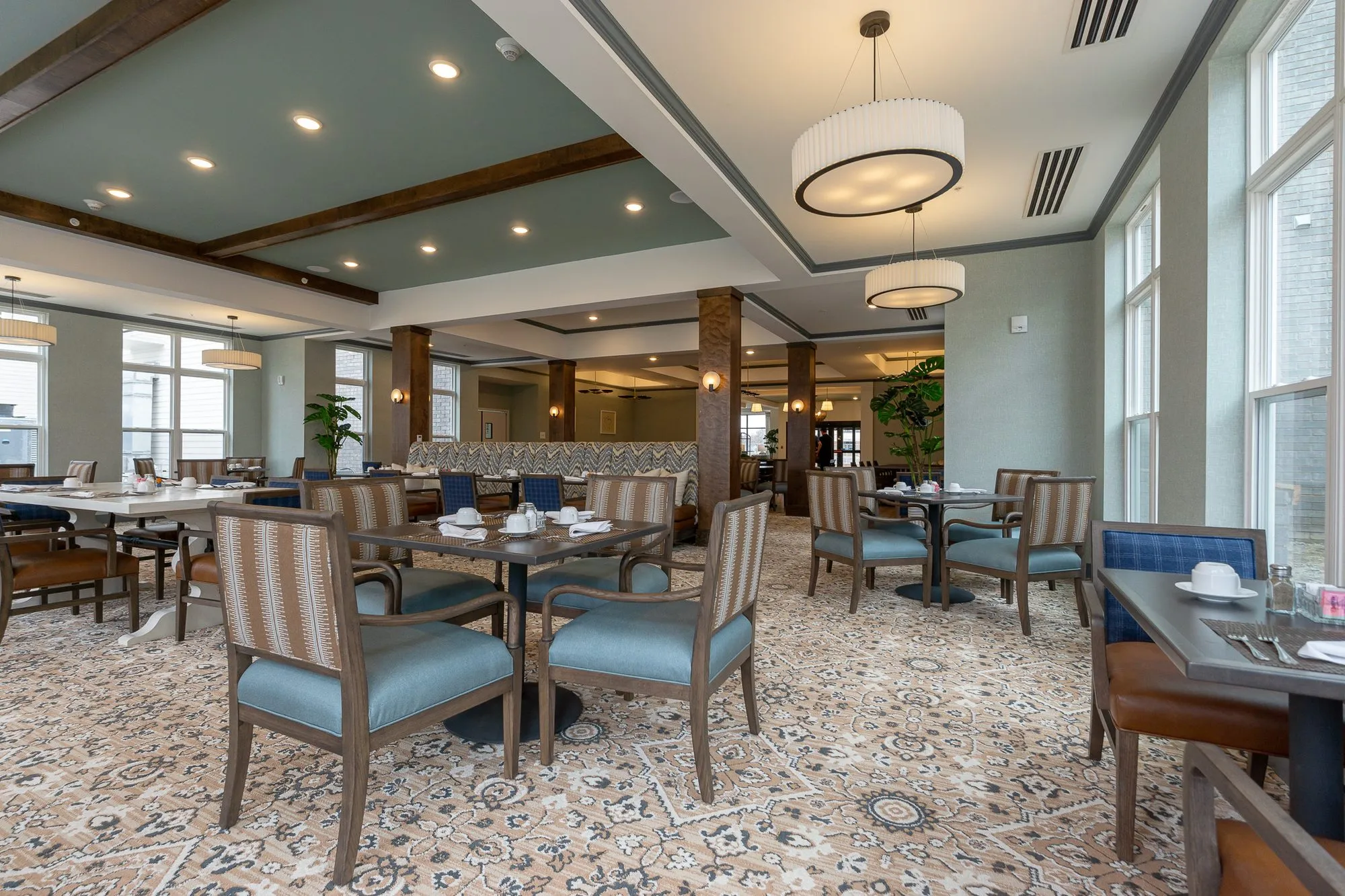Senior living dining room with blue and striped chairs at set tables under drum pendants and timber beams by tall windows