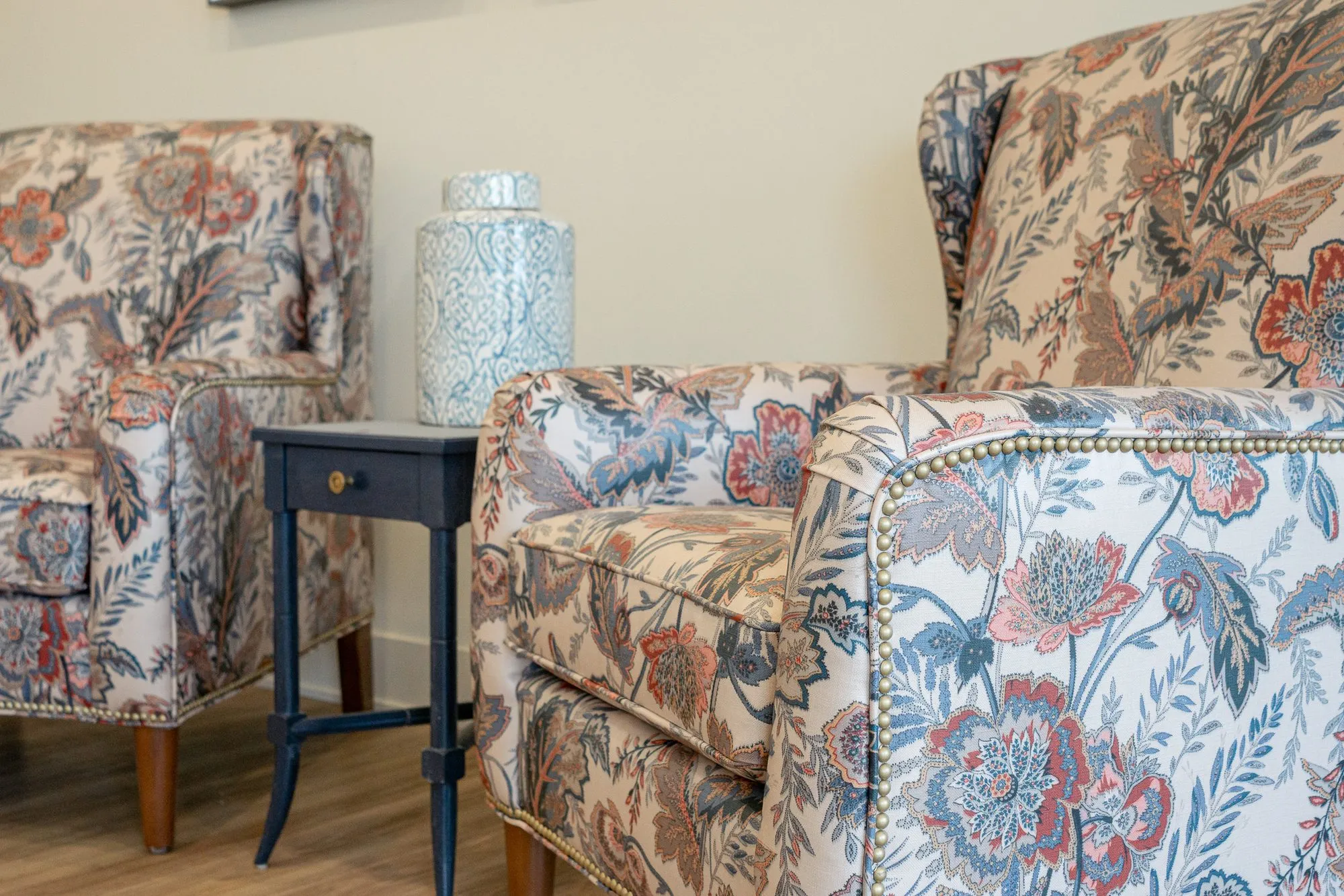 Senior living interior detail: floral wing chairs with nailhead trim beside a navy side table with a ceramic jar