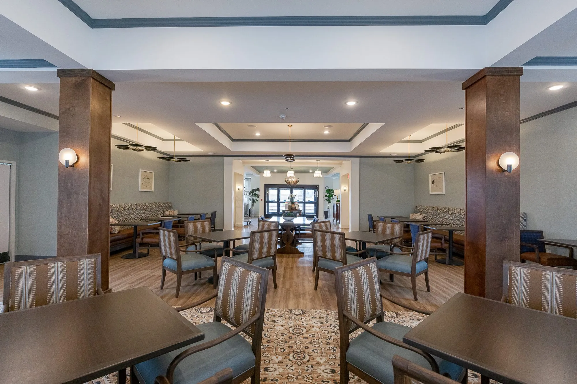 Senior living dining room framed by timber columns with sconces, striped chairs on a medallion runner to a centre table