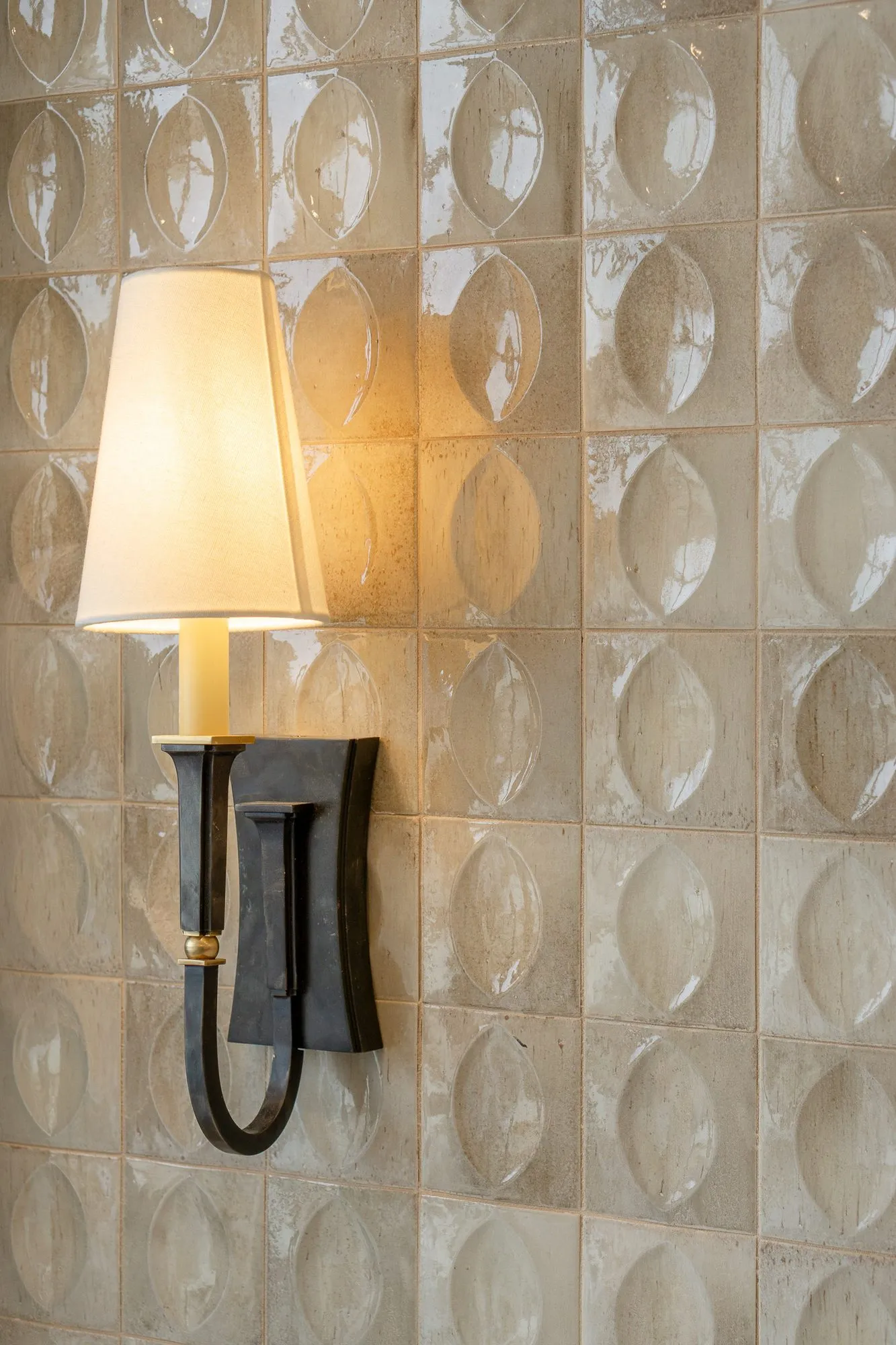 Senior living interior detail: bronze sconce glowing against glossy lens pattern tile