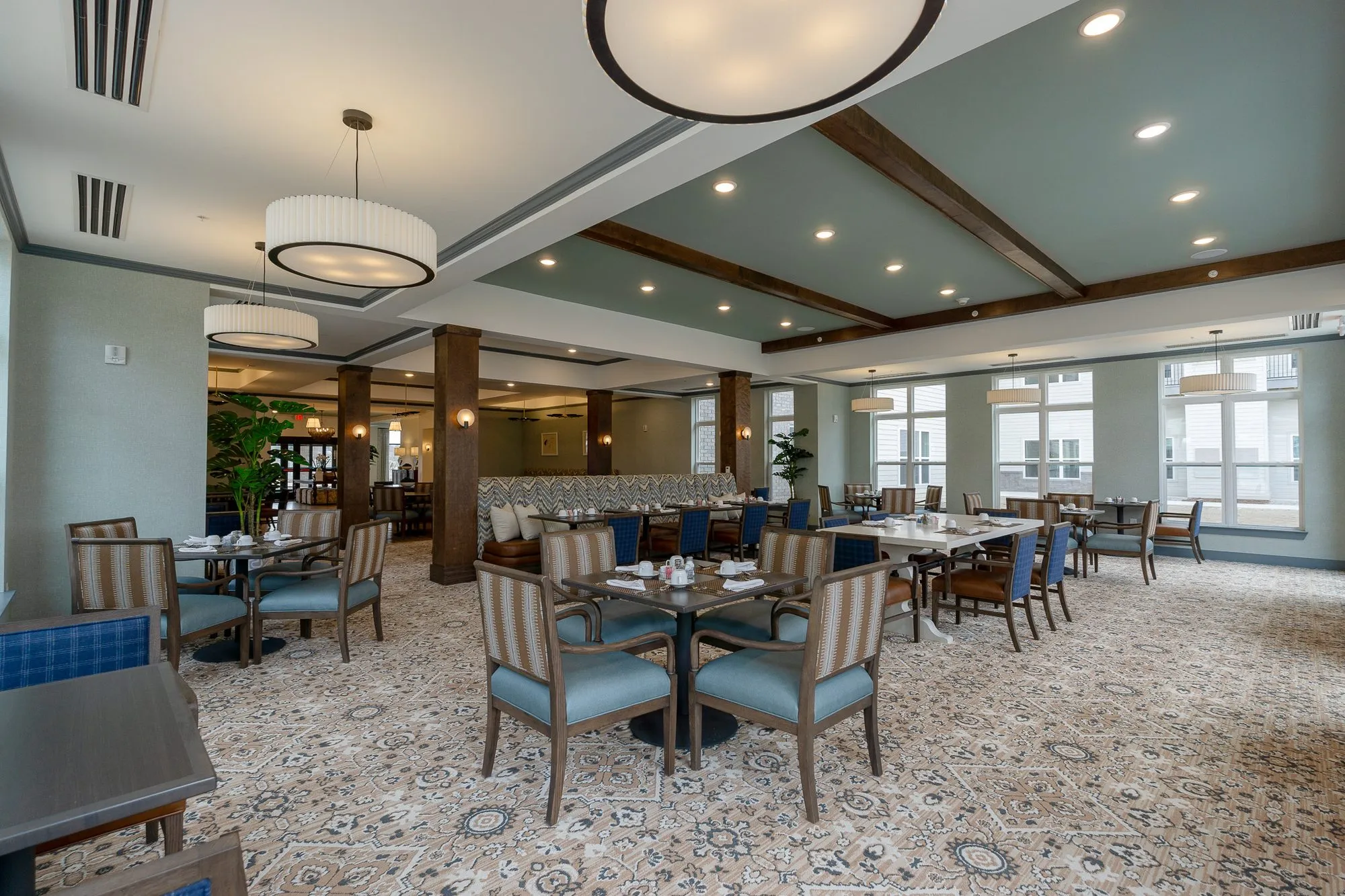 Senior living dining room under a large drum pendant, striped chairs at set tables and timber columns on a patterned carpet