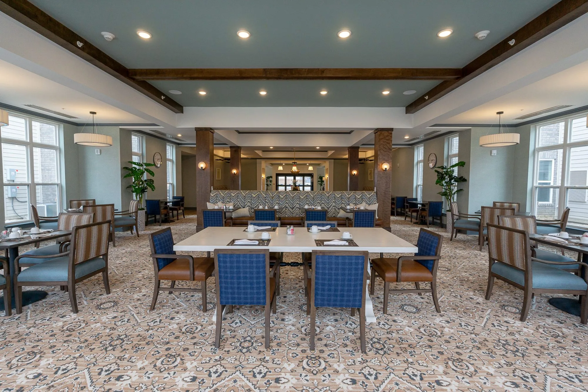 Senior living dining room in symmetry, blue plaid chairs around a long table before the chevron banquette and columns