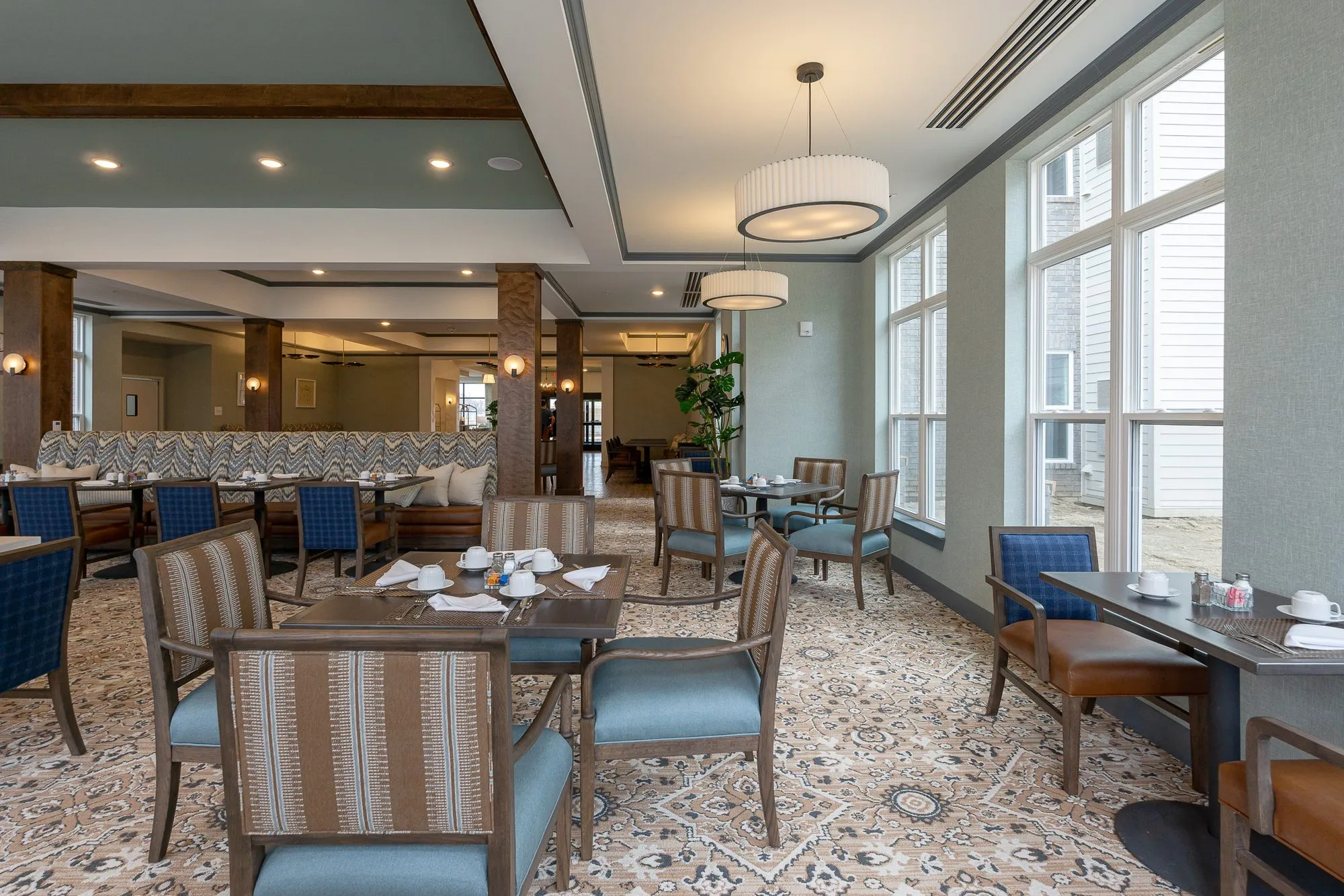 Senior living dining room with striped blue-seated chairs around a set table by tall windows, the chevron banquette beyond