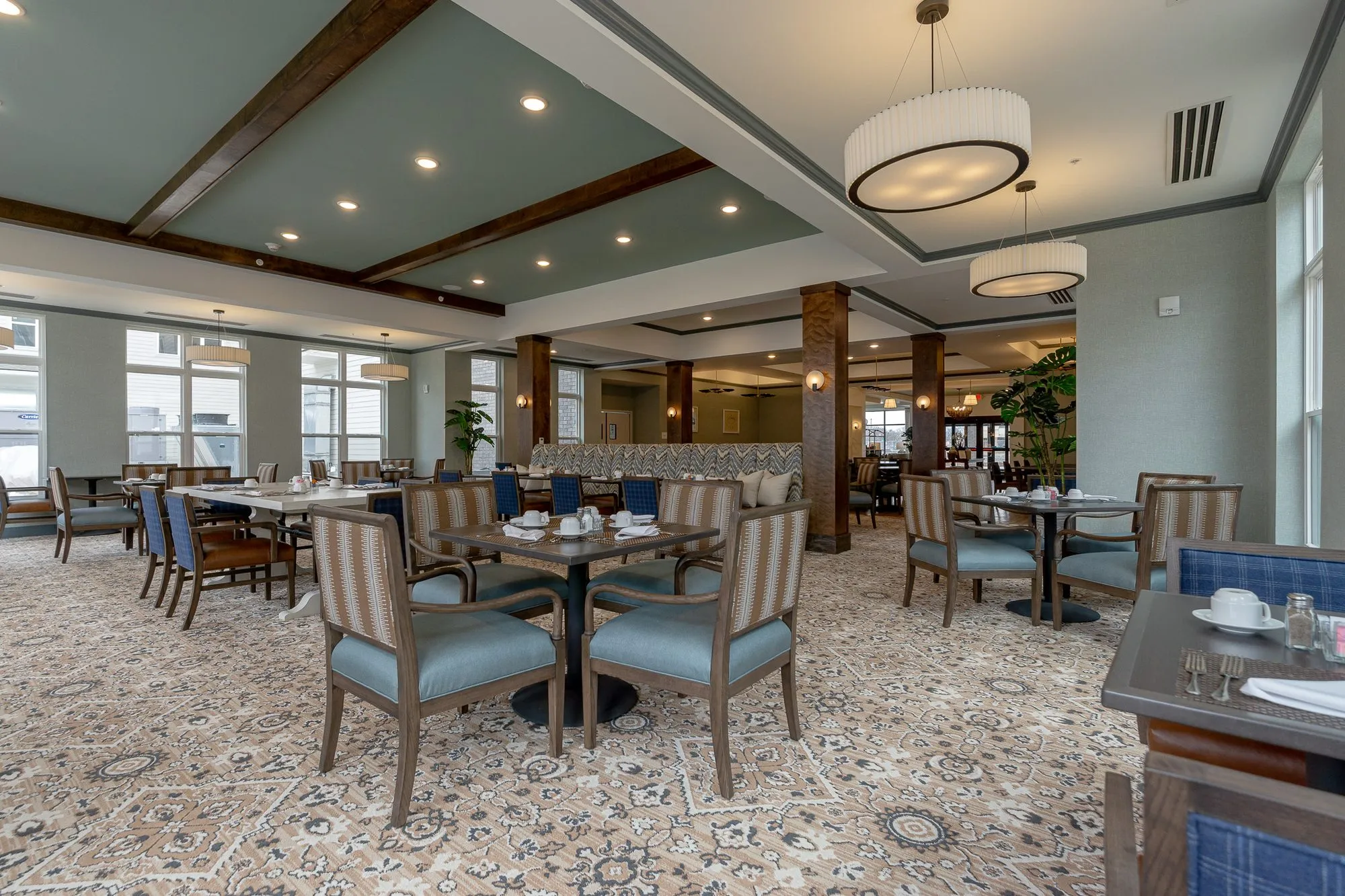 Senior living dining room with striped chairs at set tables, a blue plaid chair in the foreground and drum pendants overhead