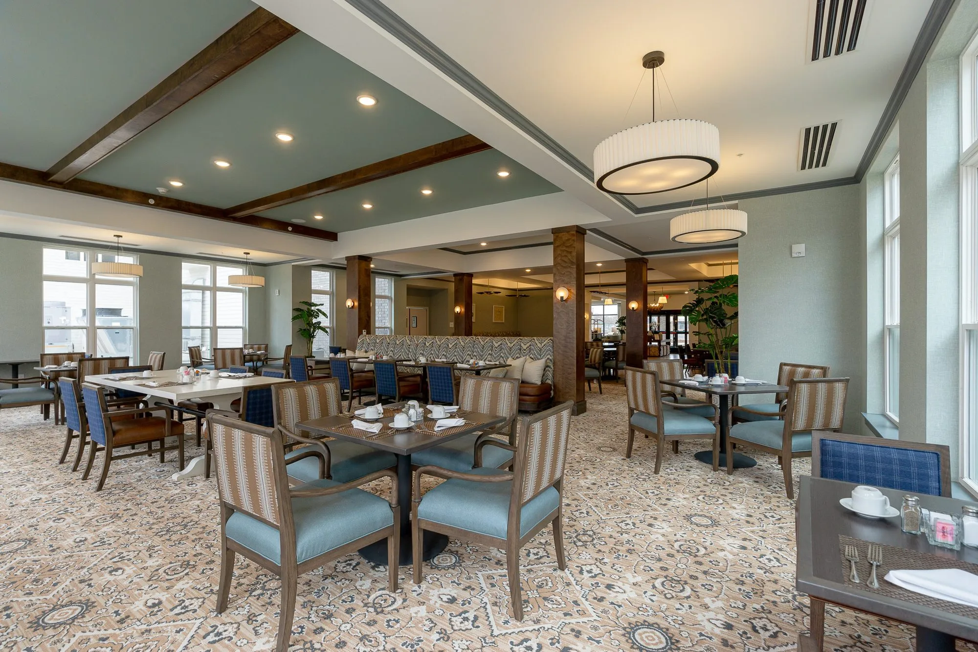 Senior living dining room under timber beams, striped chairs at set tables and drum pendants by the windows