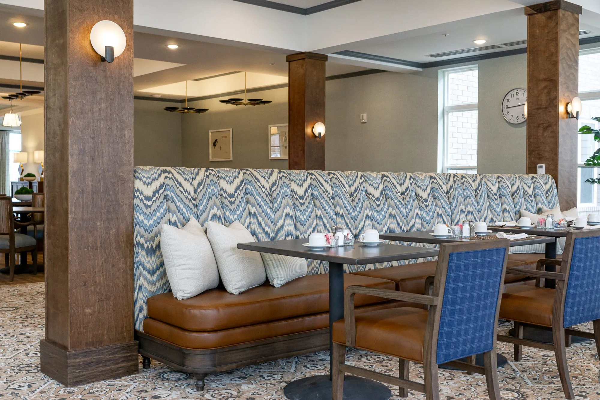 Senior living dining room with a chevron banquette and cream pillows behind blue plaid chairs at set tables, timber columns