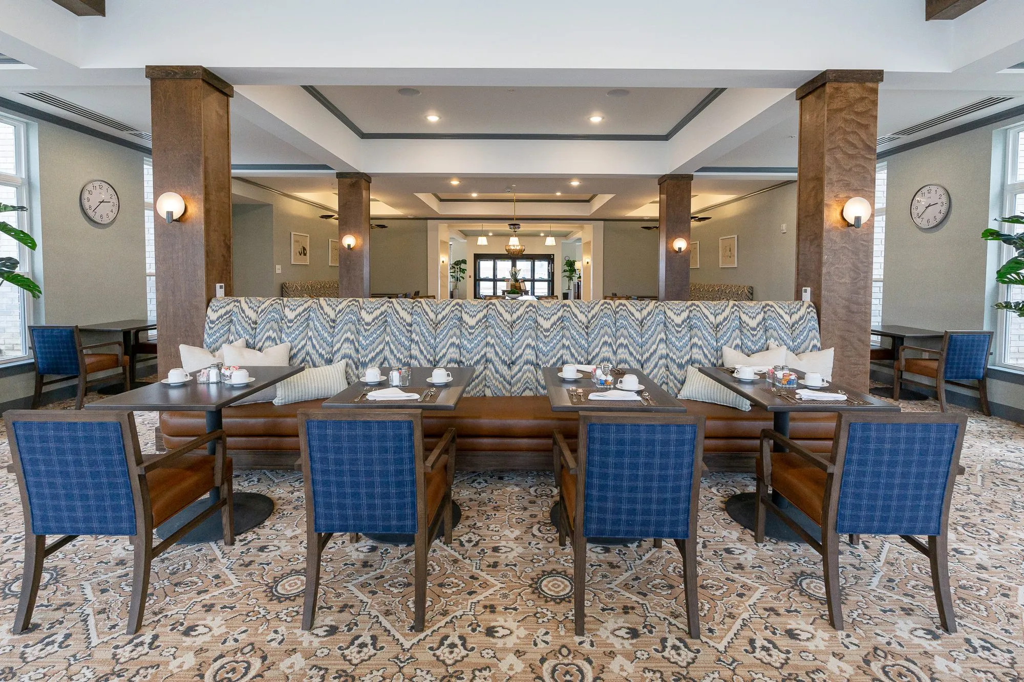 Senior living dining room with blue plaid chairs at tables before the chevron banquette, timber columns and wall clocks