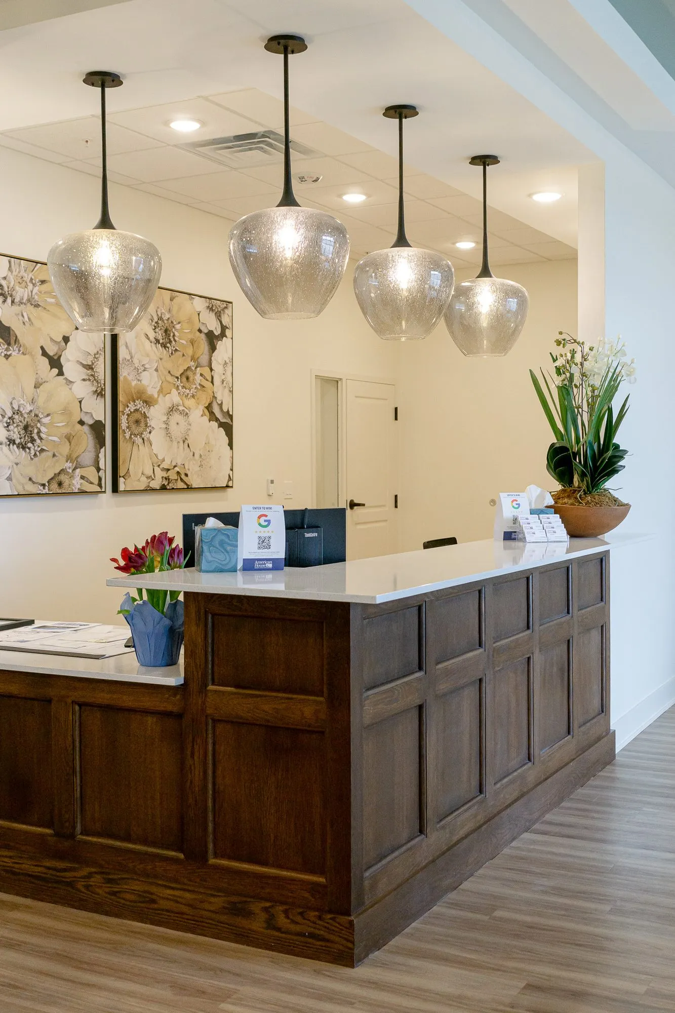 Senior living concierge desk in walnut under four smoked glass pendants beside floral canvases