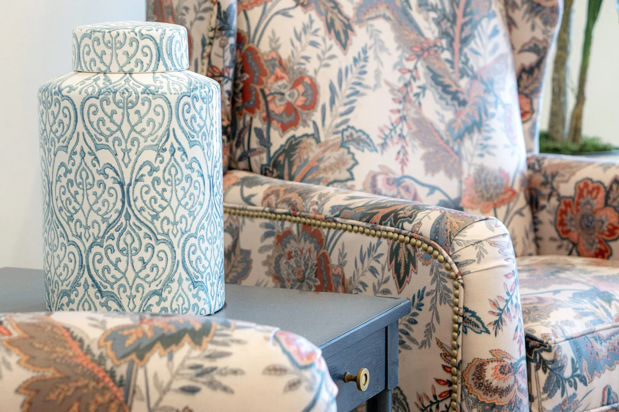 Senior living interior detail: blue and white ceramic jar on a navy table beside a floral wing chair with nailhead trim