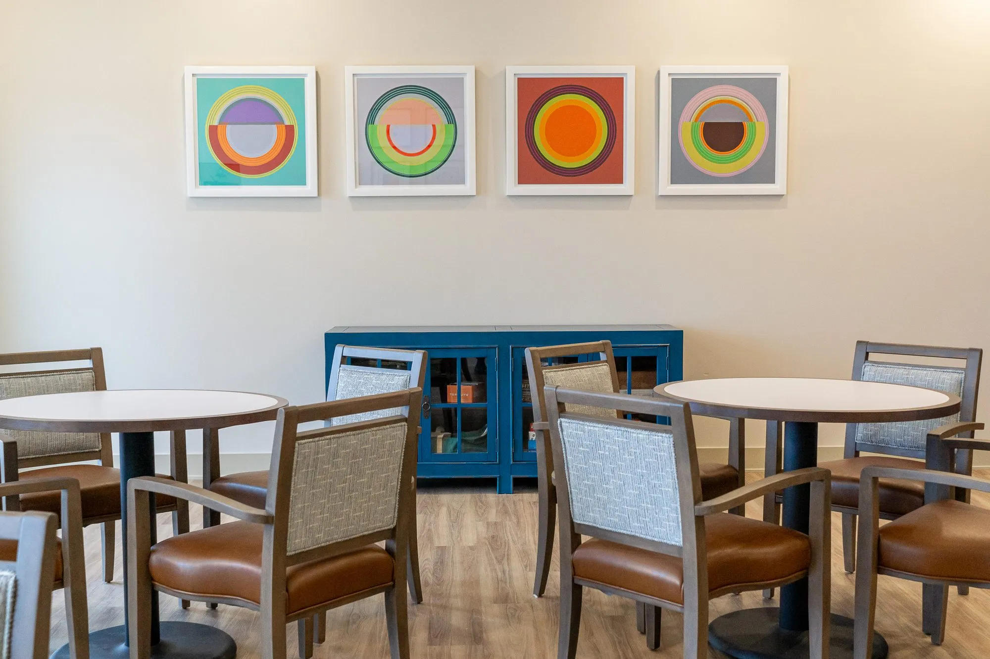 Senior living interior detail: four framed circle prints above a blue cabinet, grey wood chairs with tan seats at tables