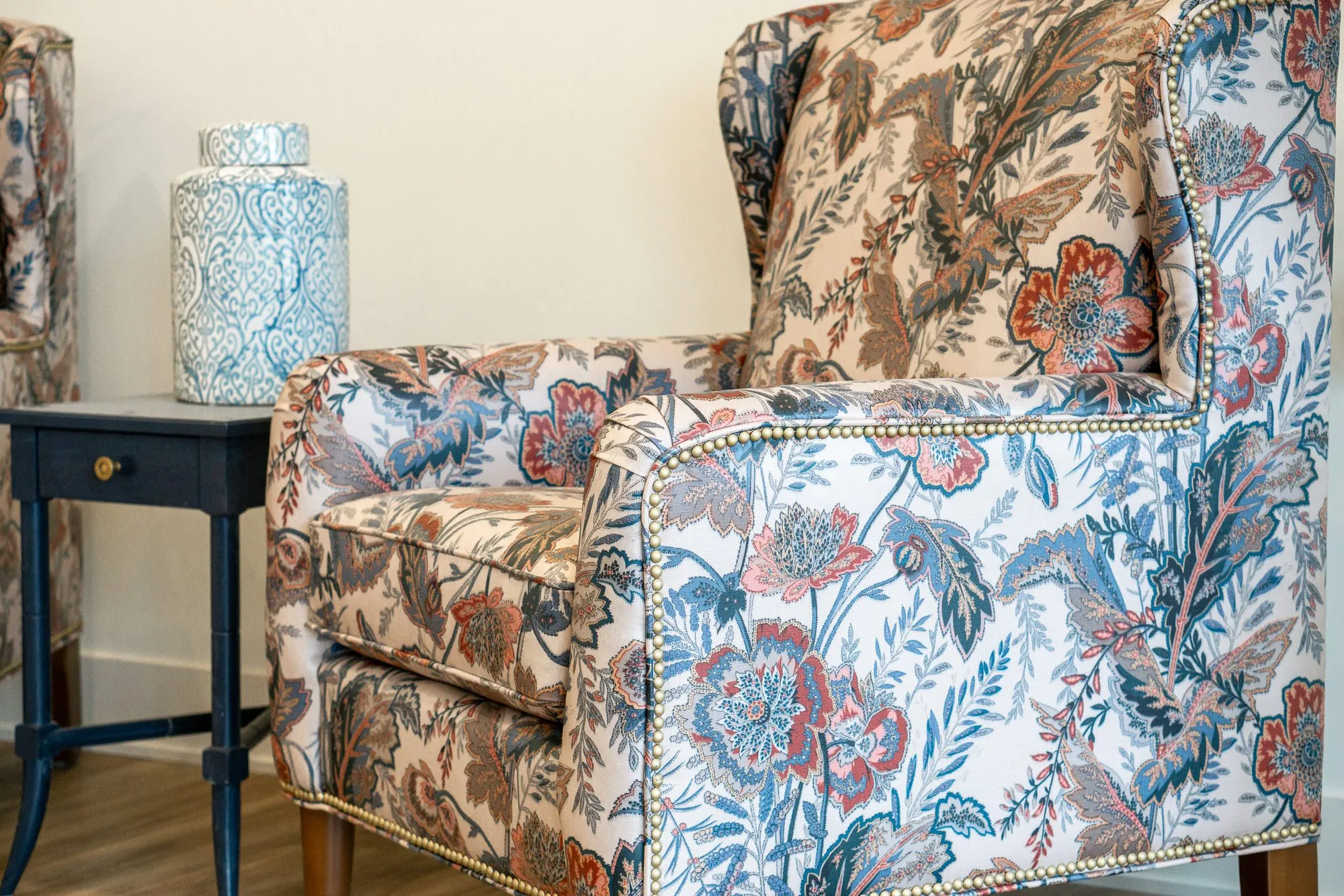 Senior living interior detail: floral print wing chair with nailhead trim beside a navy side table and a ceramic jar