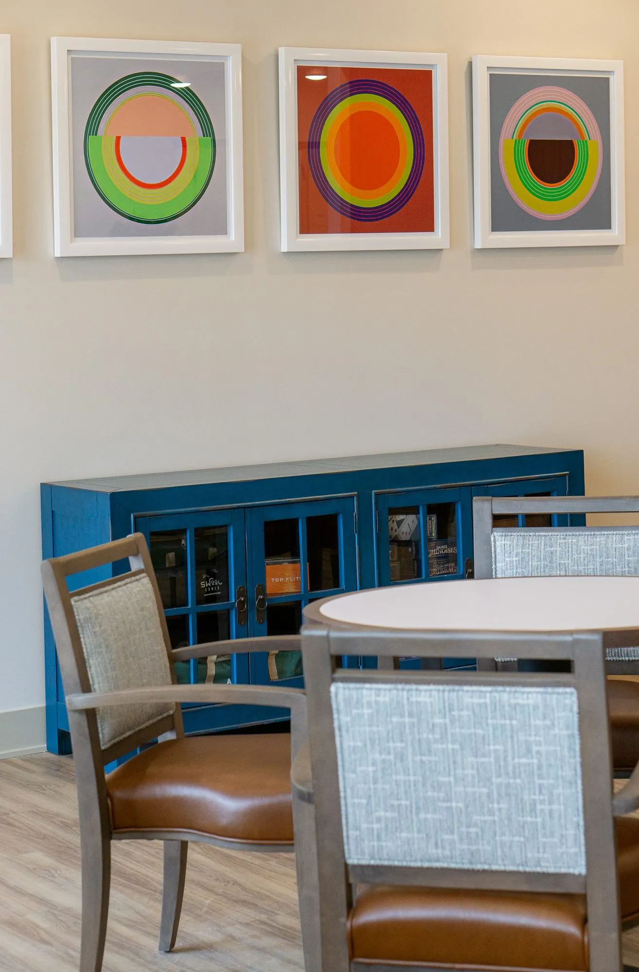Senior living interior detail: circle prints above a blue glass front cabinet, a grey wood chair at a round table