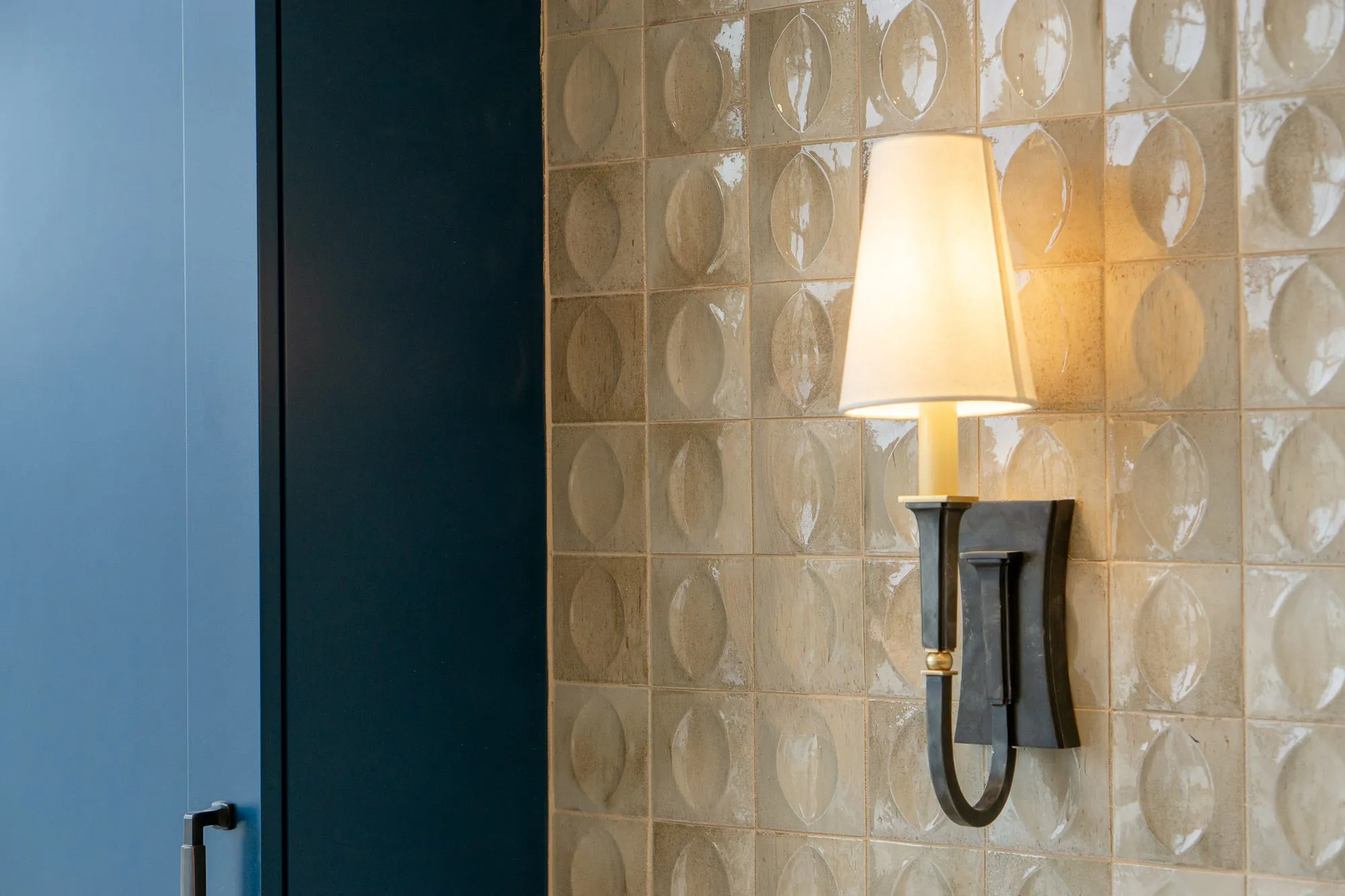 Senior living interior detail: bronze sconce on lens pattern tile, a navy cabinet edge at left