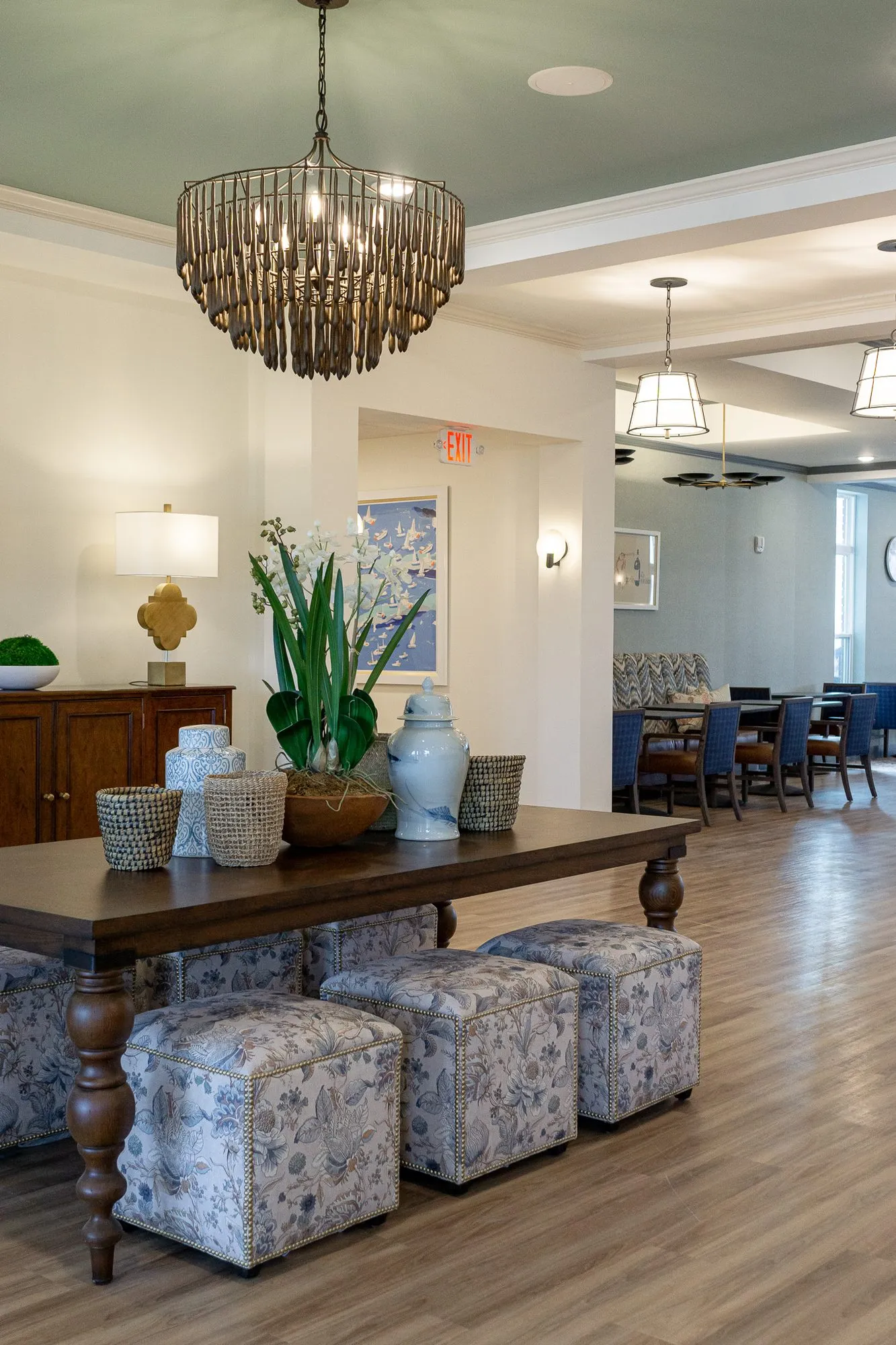 Senior living grand corridor with a walnut table and floral ottomans under a bead chandelier, a sideboard, dining room beyond