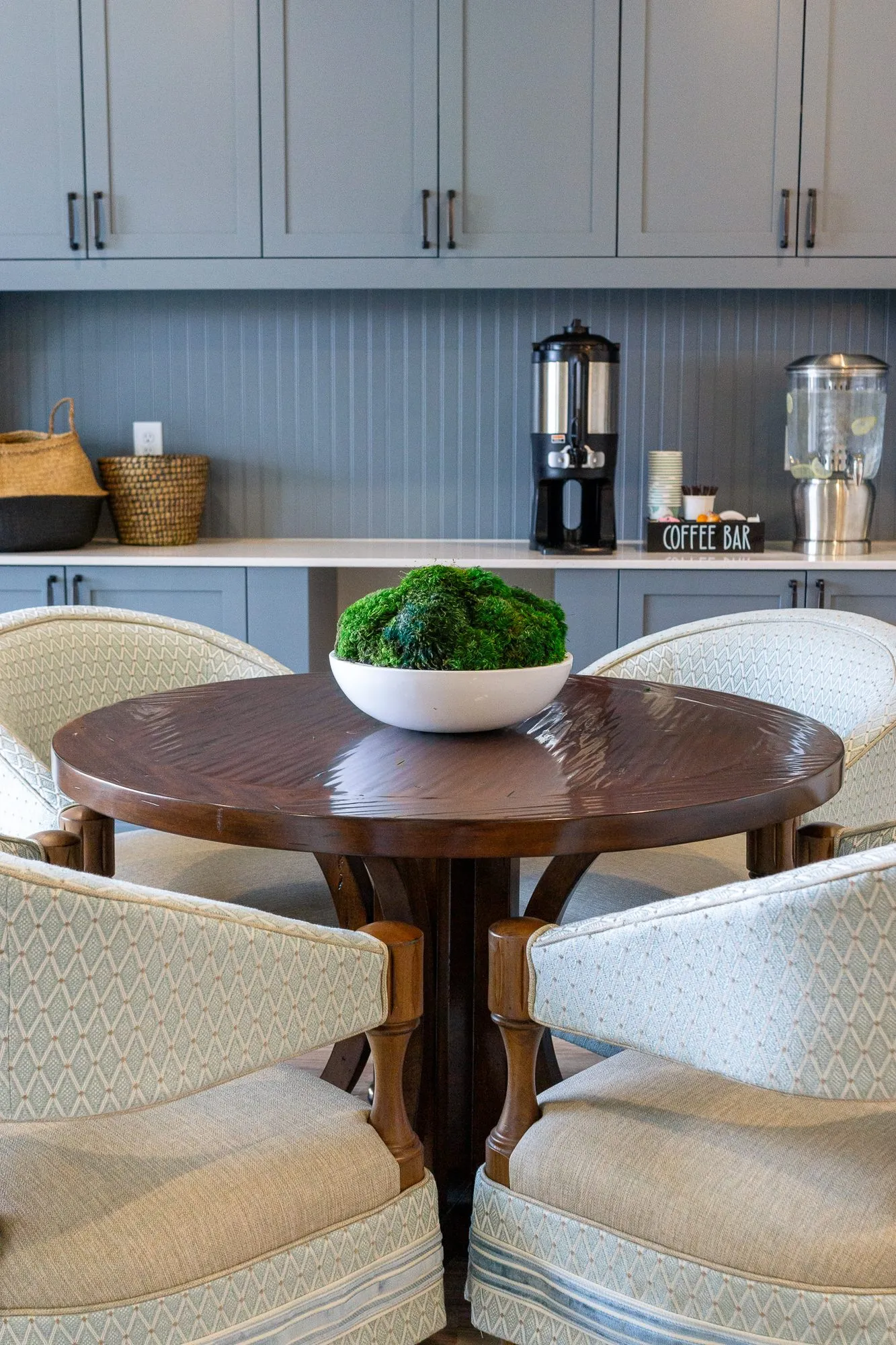 Senior living coffee bar with cream barrel chairs at a round walnut table with a moss bowl before the coffee station