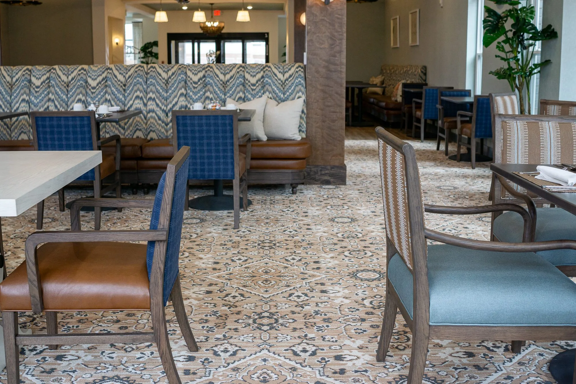 Senior living dining room with blue plaid and striped chairs at tables, the chevron banquette and a potted palm beyond