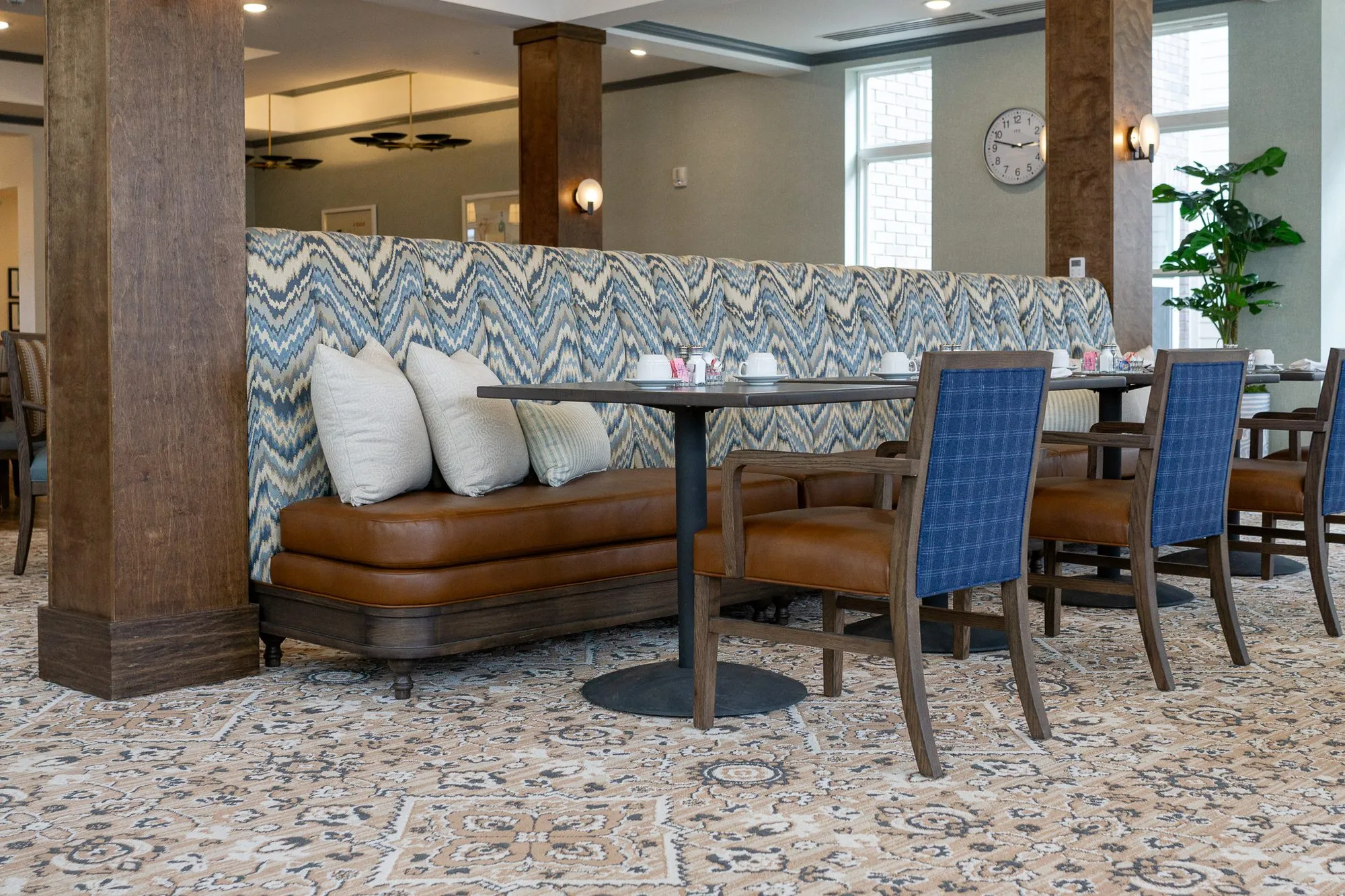 Senior living dining room with a chevron banquette, cream pillows and blue plaid chairs between timber columns, a wall clock