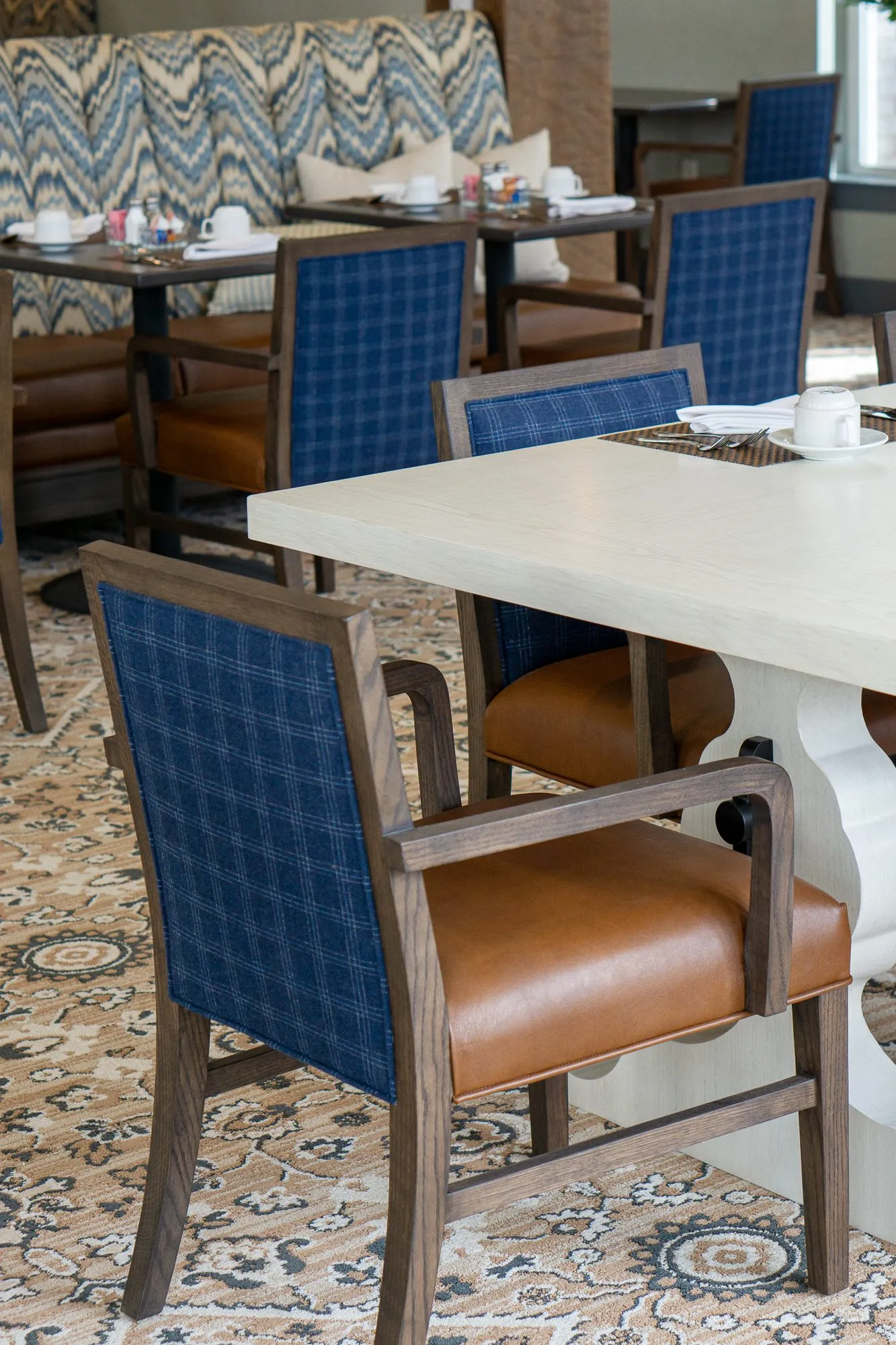 Senior living dining room with blue plaid chairs with tan leather seats at a white trestle table, the banquette beyond