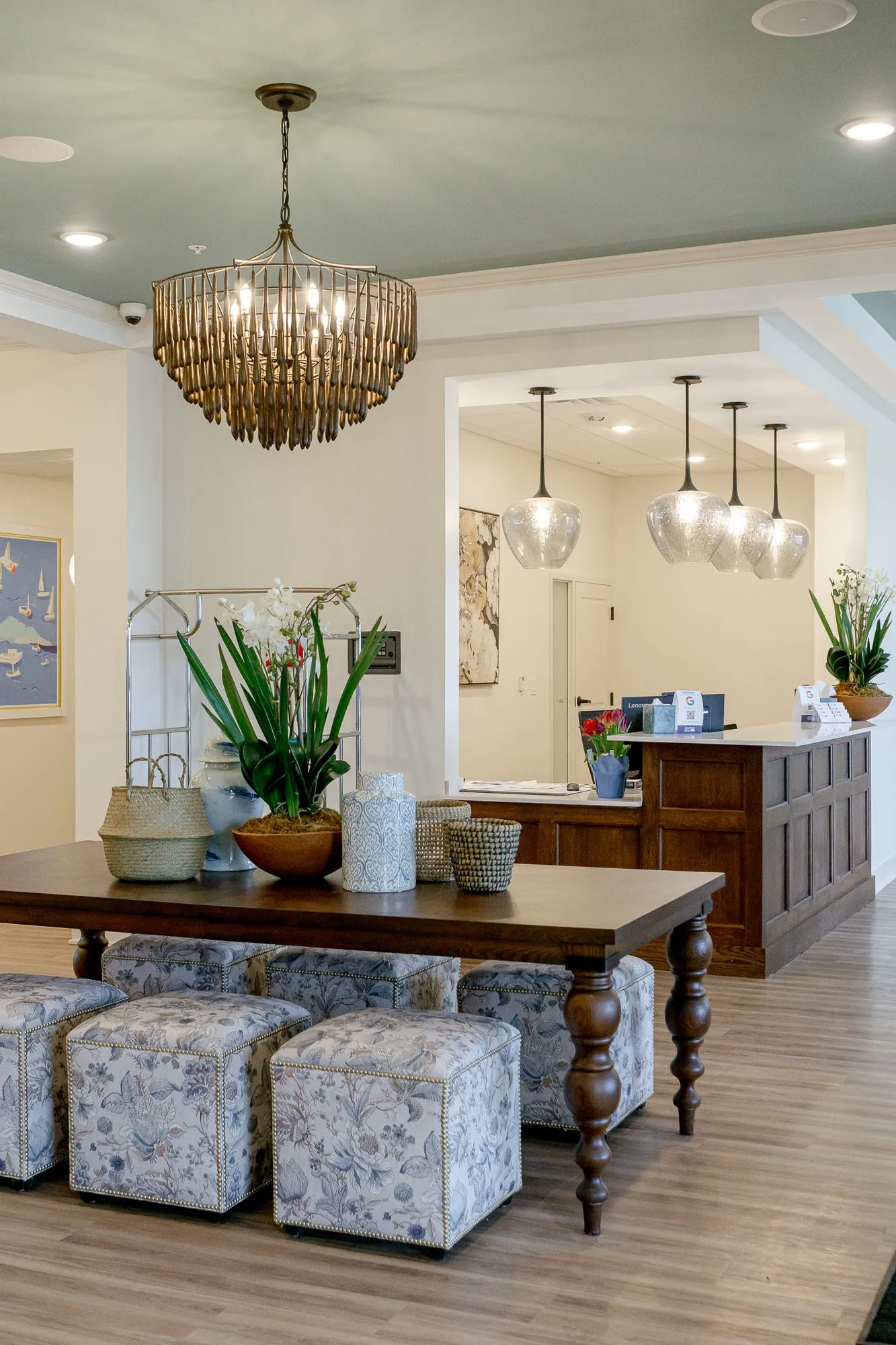 Senior living lobby with a walnut table and floral ottomans under a bead chandelier, the concierge desk and pendants beyond