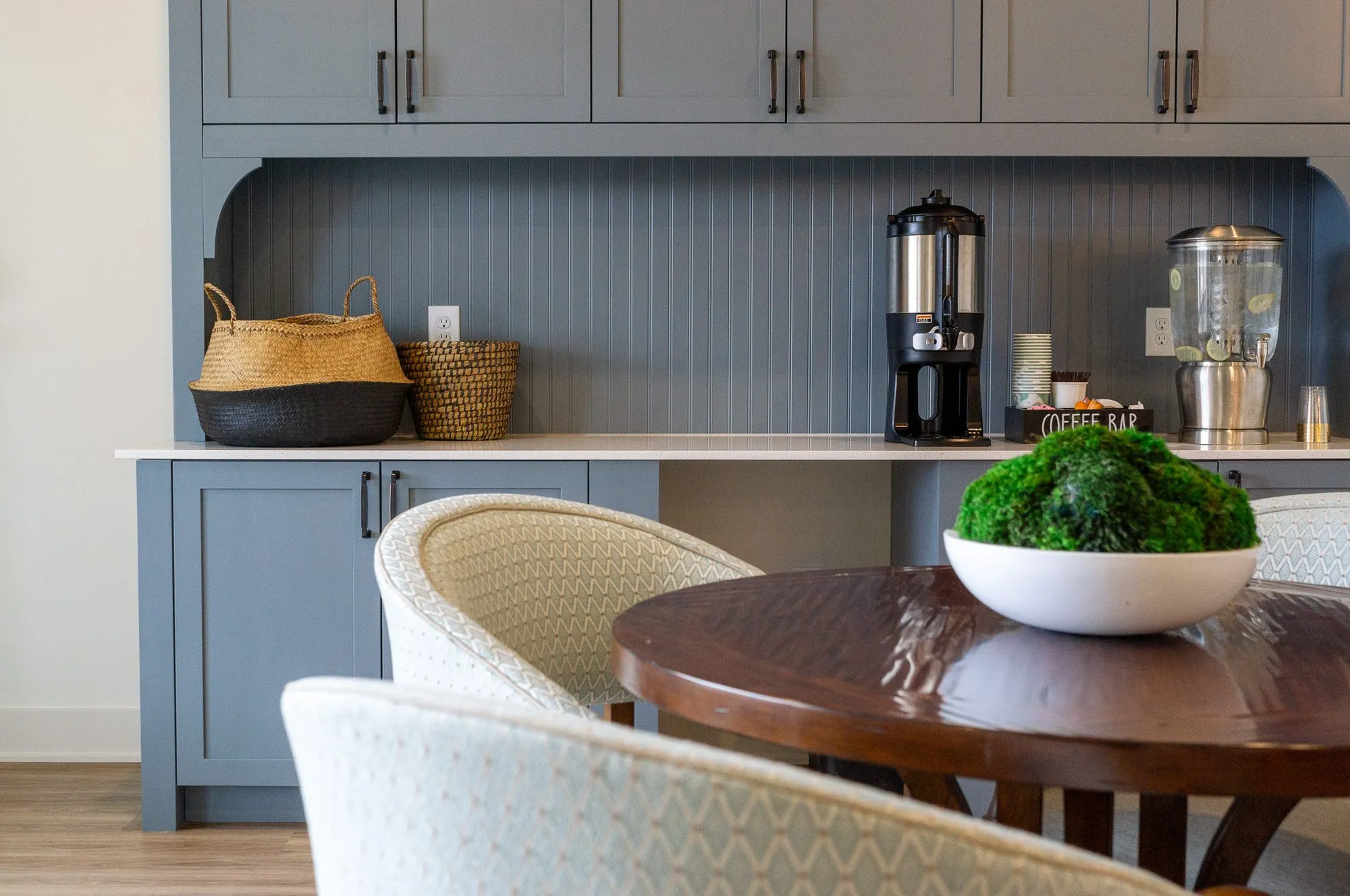 Senior living coffee station in grey-blue with a beadboard backsplash and dispensers, a moss bowl on a walnut table