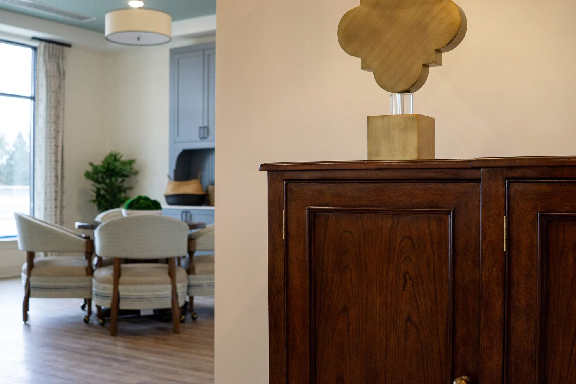 Senior living interior detail: brass quatrefoil lamp on a walnut cabinet, the coffee lounge with barrel chairs beyond