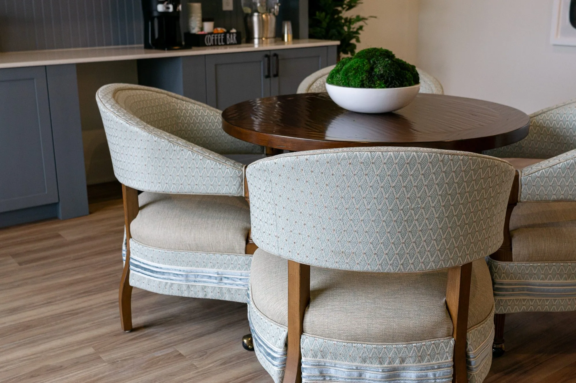Senior living coffee bar with cream barrel chairs around a round walnut table with a moss bowl, the coffee station behind
