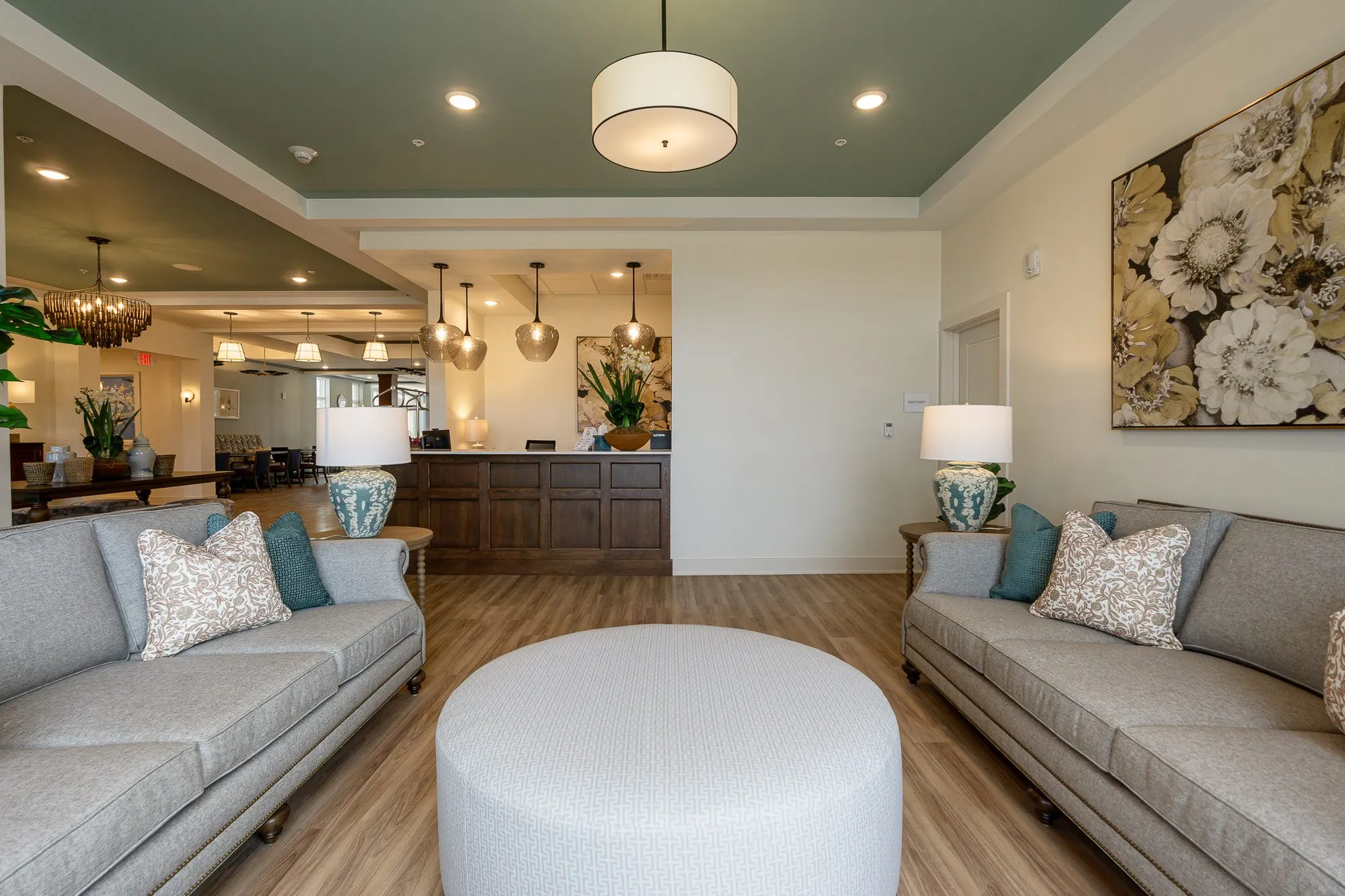 Senior living lounge with grey sofas facing across a round ottoman, the concierge desk and pendants beyond