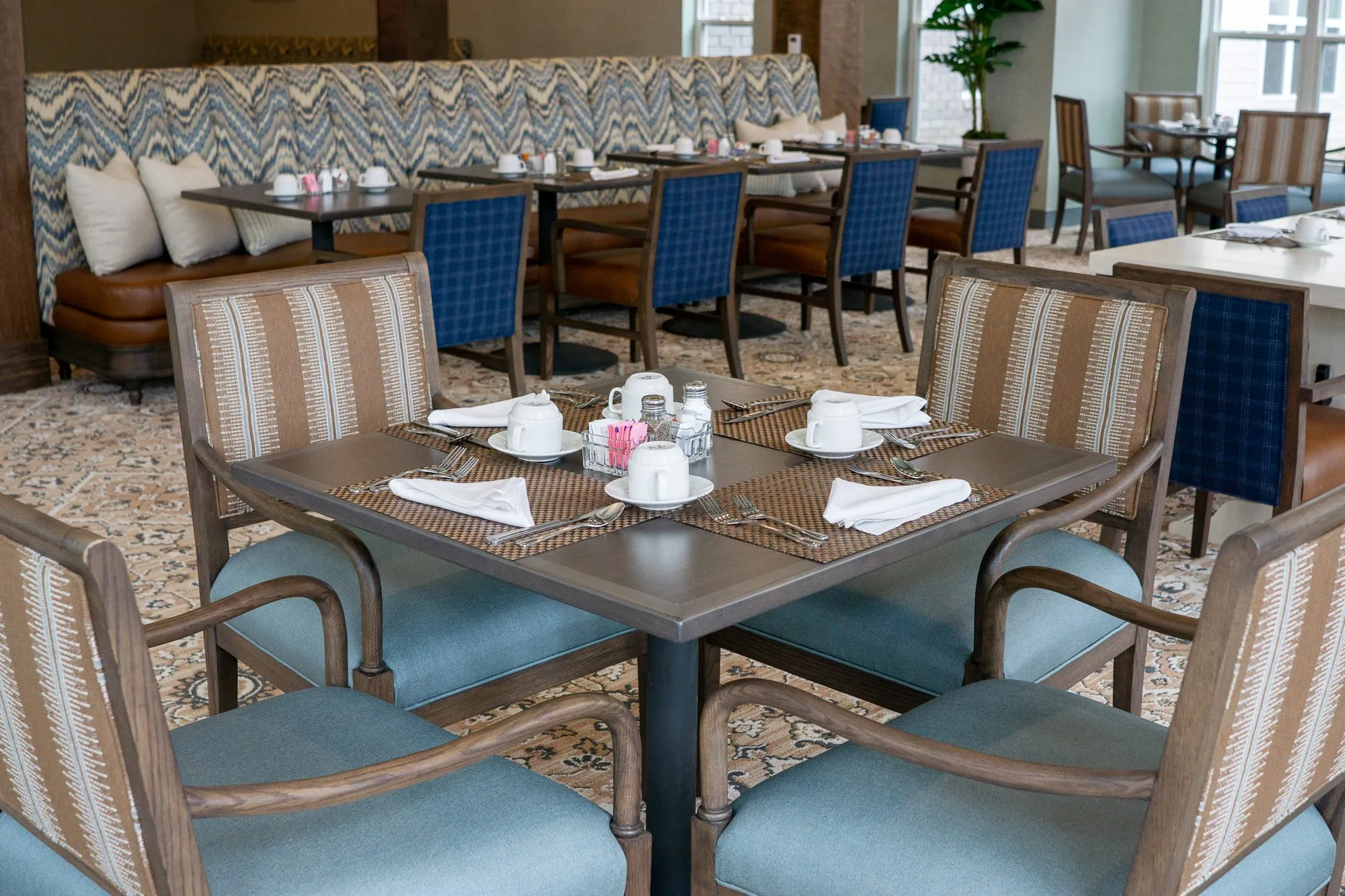 Senior living dining room with striped blue-seated chairs around a set table, blue plaid chairs and the chevron banquette