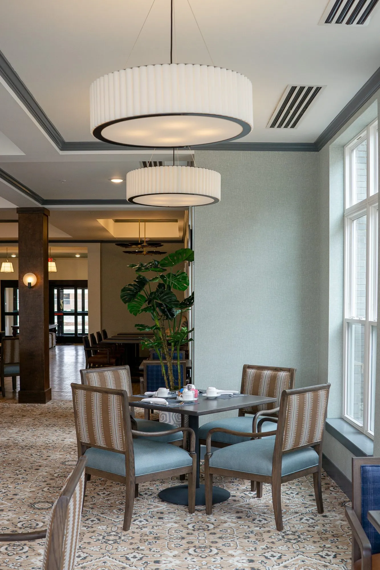 Senior living dining room with striped chairs around a table under pleated drum pendants, a potted plant by the window