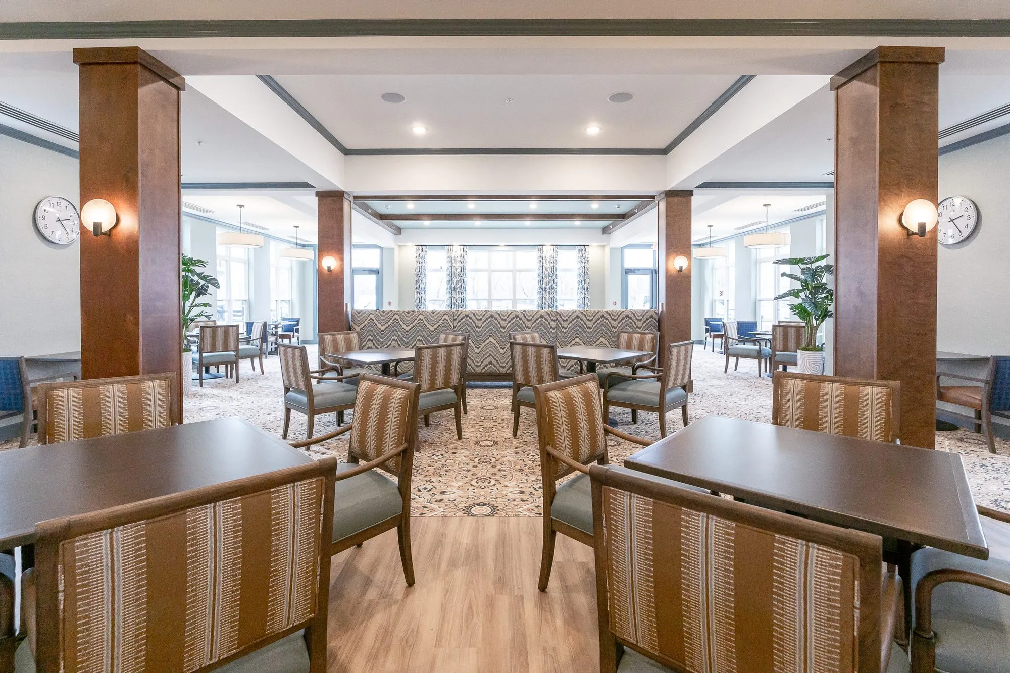 Senior living dining room with striped chairs at tables between timber columns with sconces and wall clocks