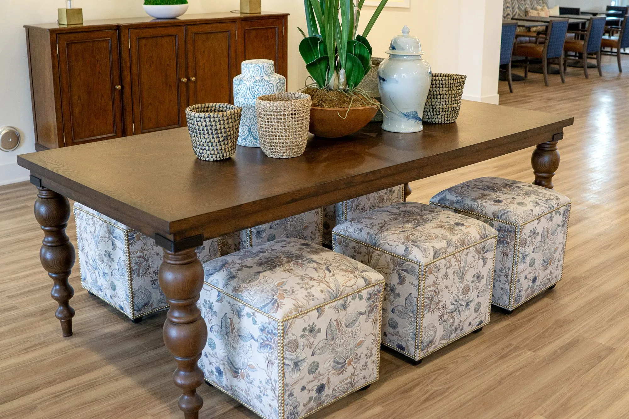 Senior living interior detail: walnut table with woven baskets, ceramic jars and floral cube ottomans, a sideboard behind