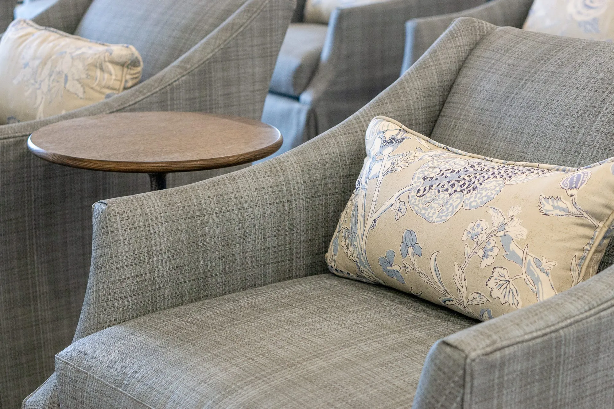 Senior living interior detail: grey plaid armchair with a floral pillow beside a round drink table