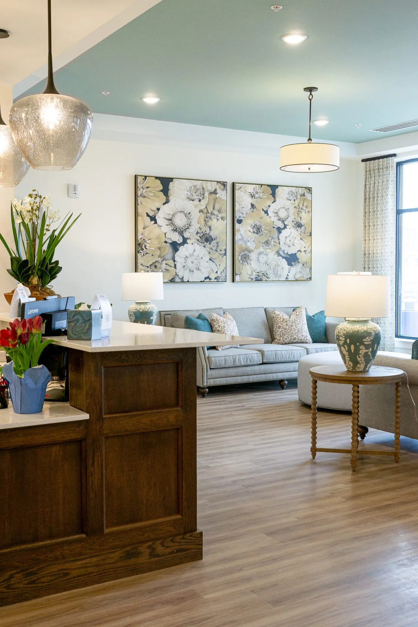 Senior living lounge seen past the concierge desk, a grey sofa below two floral canvases