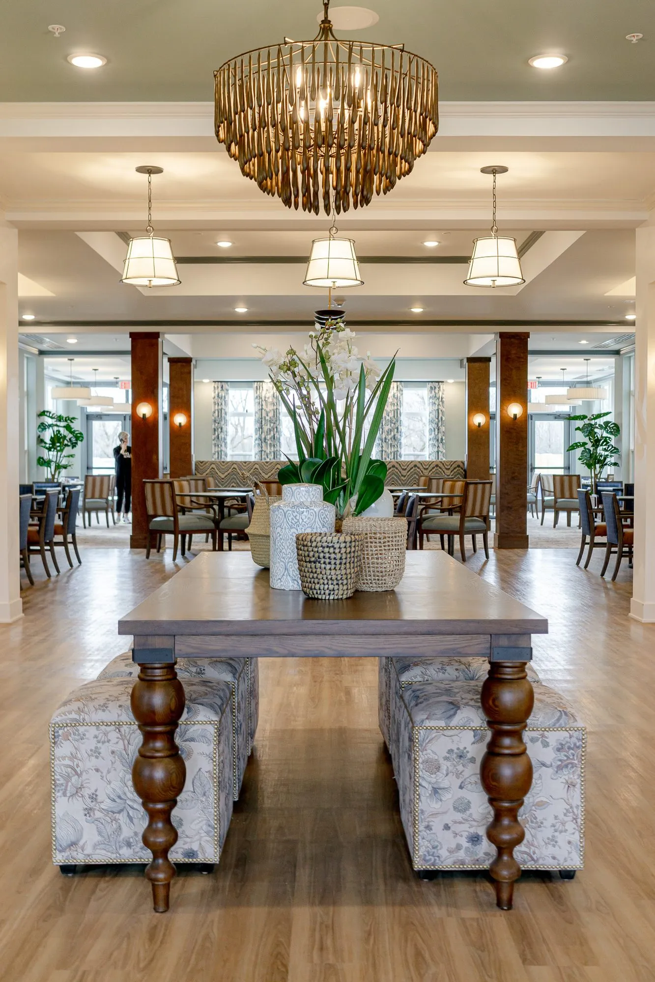 Senior living grand corridor with a walnut table and floral ottomans under a bead chandelier, the dining room and its columns