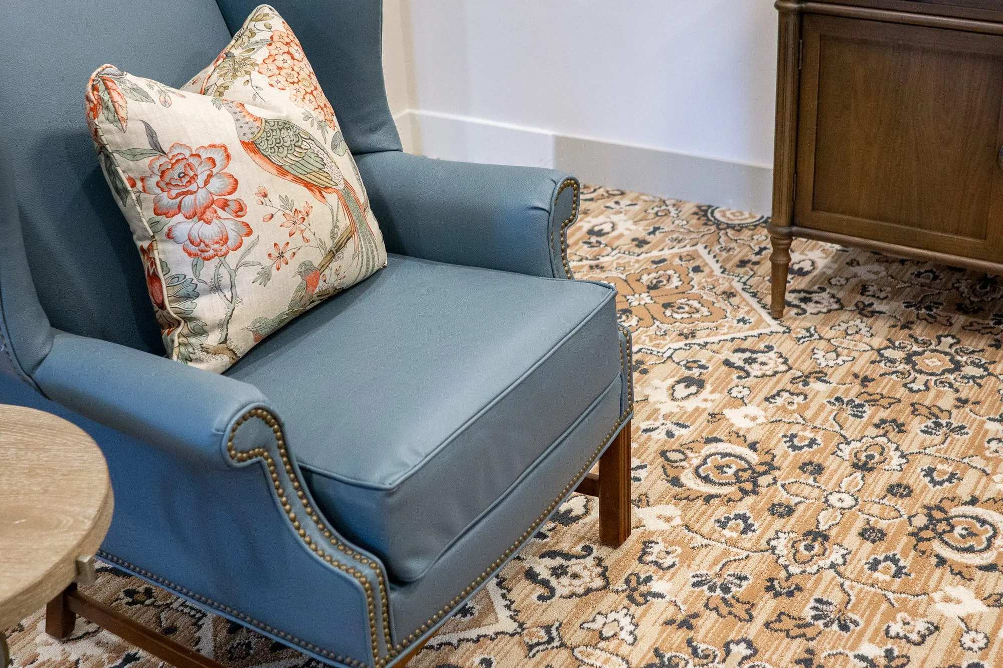 Senior living interior detail: blue leather wing chair with a bird and floral pillow beside a walnut cabinet