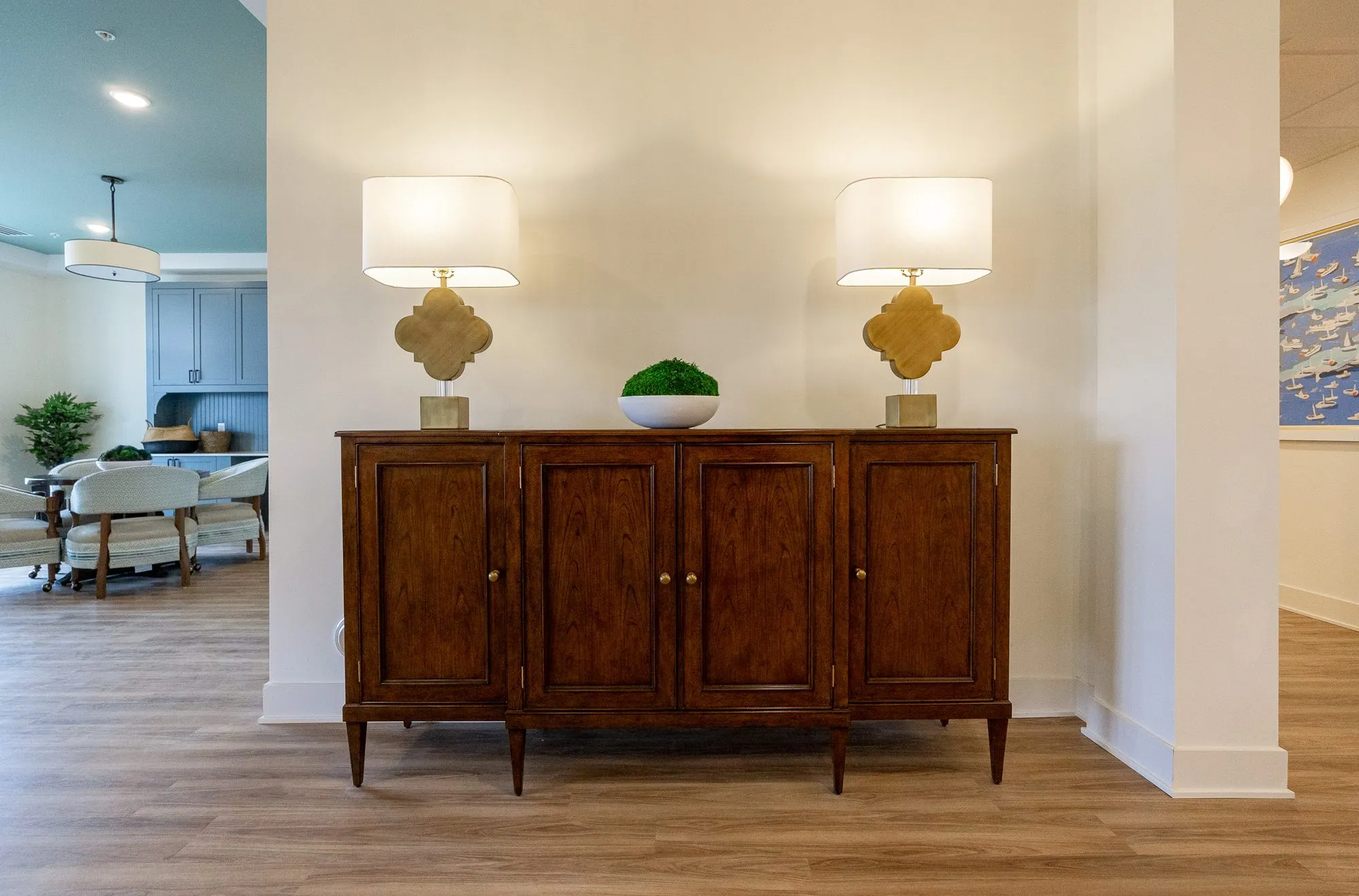 Senior living interior detail: walnut sideboard with two brass quatrefoil lamps and a moss bowl, the coffee lounge at left