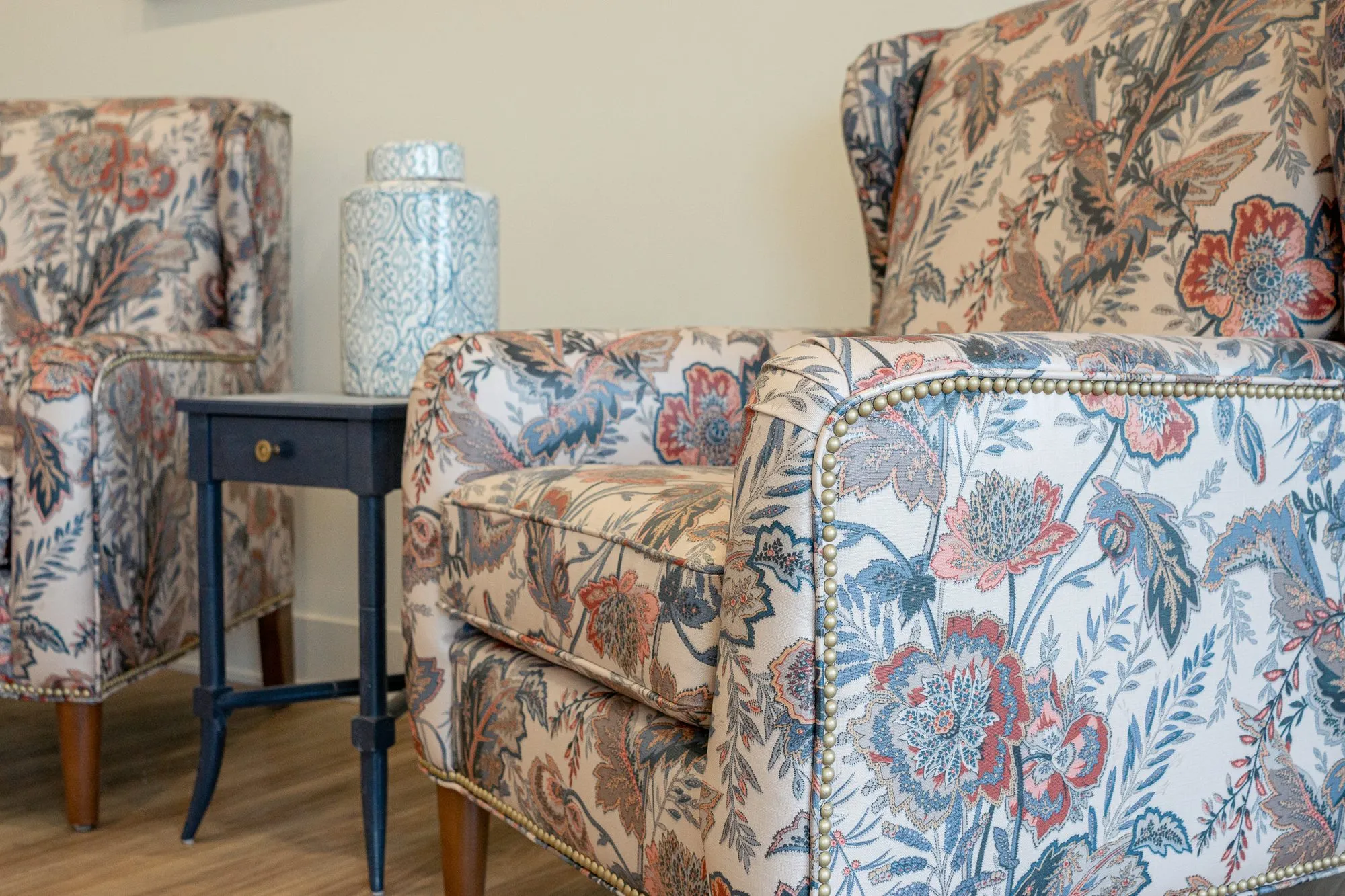 Senior living interior detail: floral wing chairs with nailhead trim flanking a navy side table with a ceramic jar